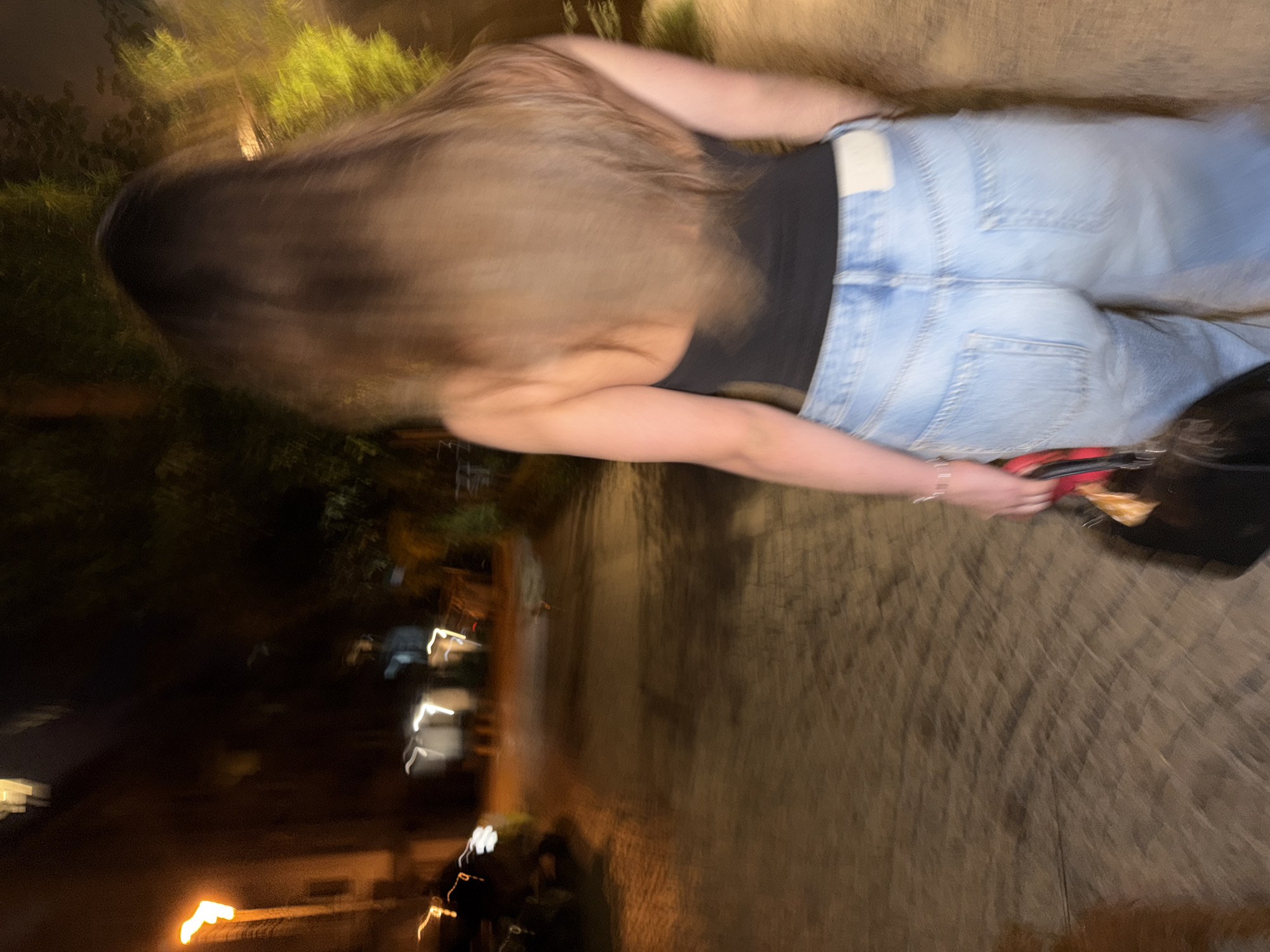 The image depicts a person walking away from the camera down a paved path at night. The person is wearing a black top and light blue jeans. They are carrying a black bag with a red strap. The background shows trees and buildings illuminated by streetlights, suggesting an urban environment. The scene is set in Sofia, Bulgaria, near the National Palace of Culture. The motion blur suggests the photo was taken while the person was in motion.