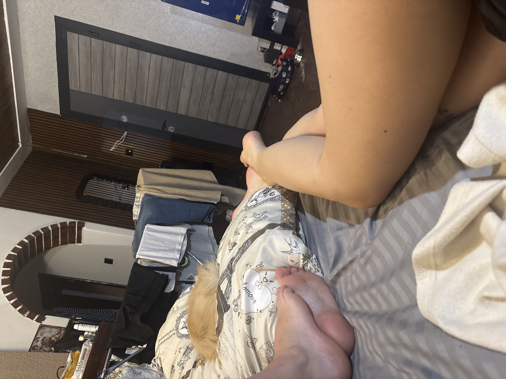 A close-up shot inside a room shows a person's legs, feet, and part of their torso on a bed. The bedding is striped grey. The person has fair skin. Part of a decorative pillow or blanket with a print, including the word "amour," is visible. In the background, there is a partially visible doorway or entrance, with a textured wall and a glimpse of a shelf. There are some clothes stacked up in the background. The location is a room, not explicitly identifiable as being in Belopoptsi, Bulgaria, from the image itself.