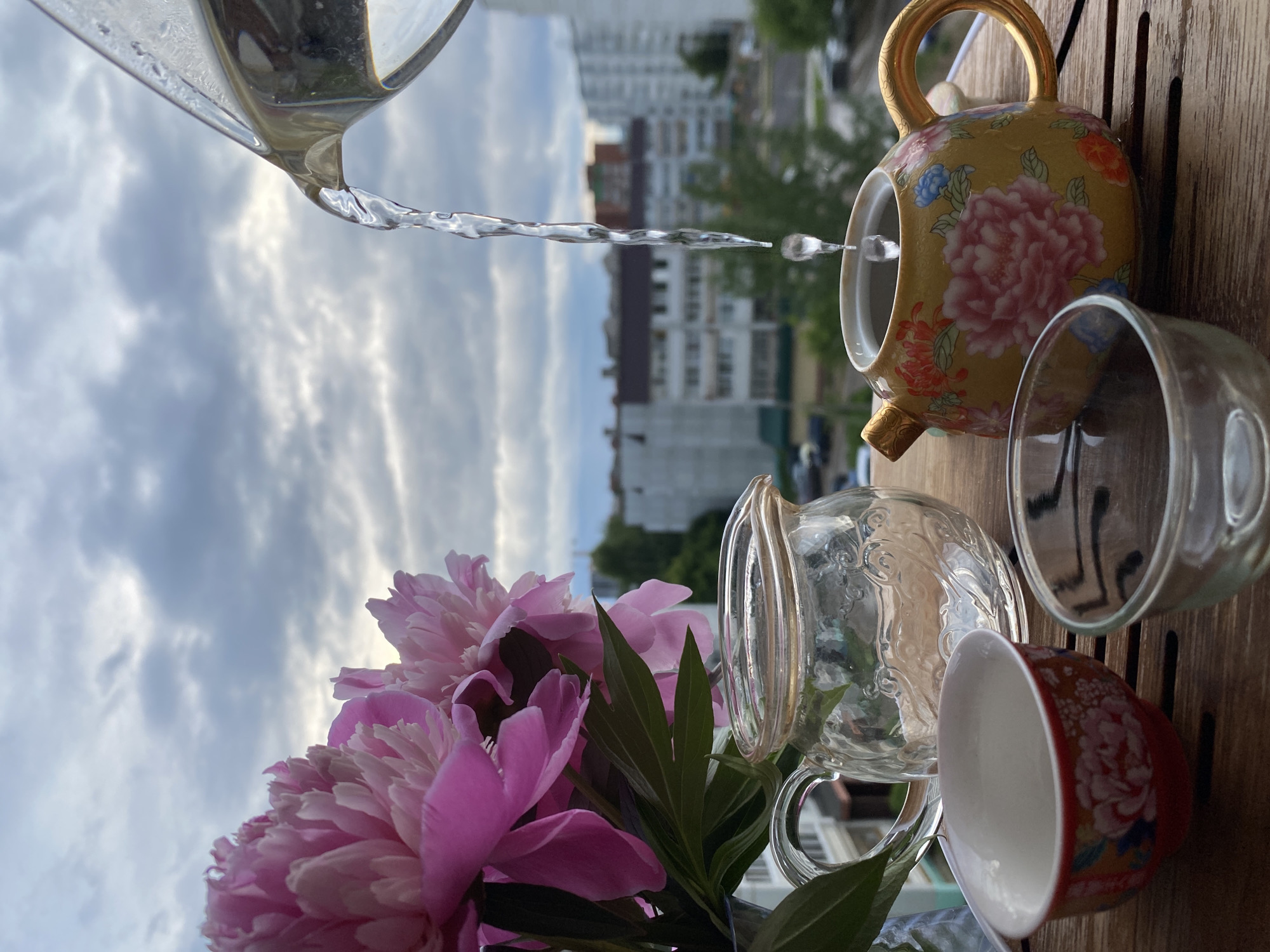 The image captures an outdoor still life scene situated on a wooden slatted surface in the city of Chaikovsky, Russia. On the left, a bouquet of vibrant pink peonies with green foliage is partially visible in a clear vase. To the right of the flowers stands an empty clear glass pitcher with an embossed patterned surface. In the lower left foreground, a small, ornate red and white teacup with a floral peony design is present. Adjacent to it on the right, a small, clear glass bowl contains a dark liquid. The central focus on the right is a decorative yellow teapot featuring detailed painted floral motifs, primarily pink peonies, with a golden handle and spout. Water is actively being poured into the teapot from an upper right, partially visible clear glass vessel, forming a distinct stream and a single suspended droplet just above the teapot's opening. The background consists of an urban landscape featuring multiple multi-story residential buildings, interspersed with green trees and bushes, under a cloudy sky composed of grey and white clouds. A street with parked cars is discernable in the mid-ground. No individuals are depicted in the scene.