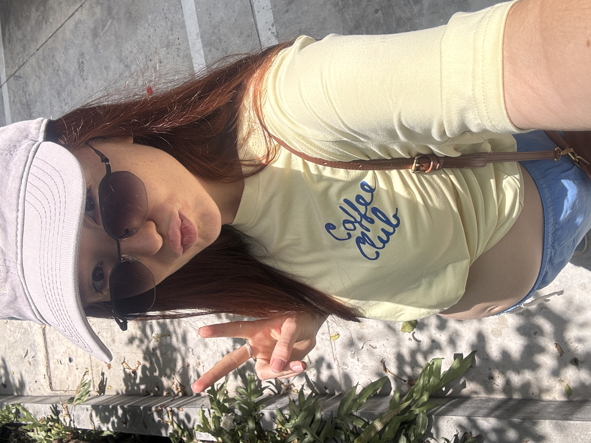 A young woman poses outdoors. She wears sunglasses, a light gray baseball cap, a pale yellow "Coffee Club" t-shirt, and blue shorts. A thin brown strap of a purse is visible across her chest. Her red hair is long. She holds up two fingers in a peace sign. She is standing near a low wall with green plants. The location is not identifiable as Choeng Thale, Thailand.