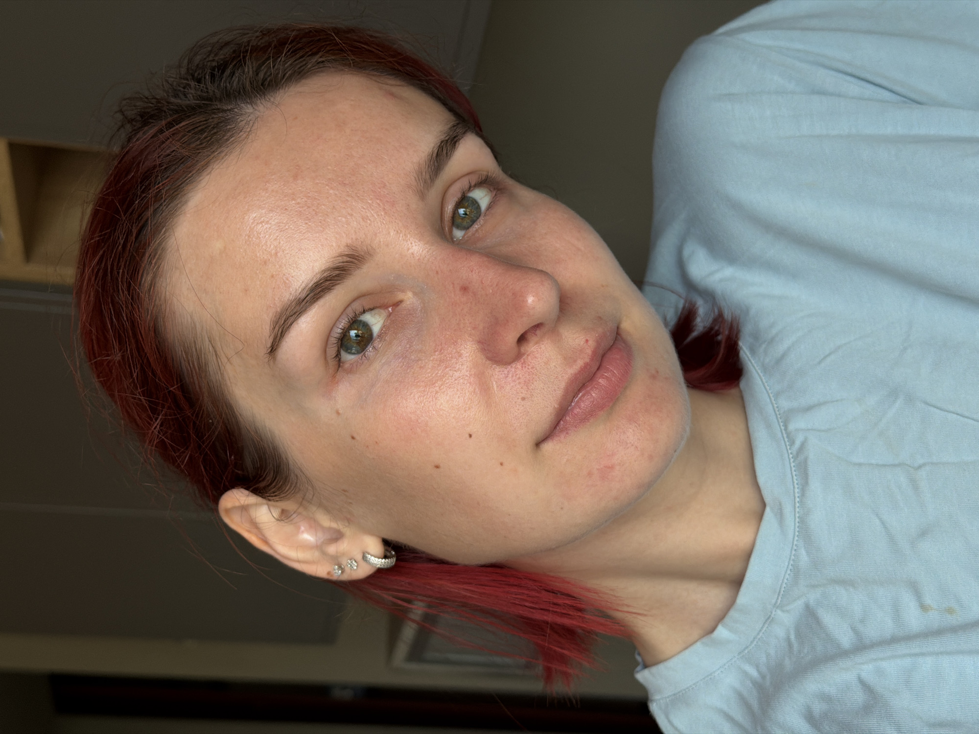 A close-up portrait of a woman with reddish-brown hair, light skin, and green eyes, wearing a light blue shirt. She has multiple piercings in her left ear. The background is dark and out of focus. There is no information in the image to indicate the location is Chaikovsky, Russia.
