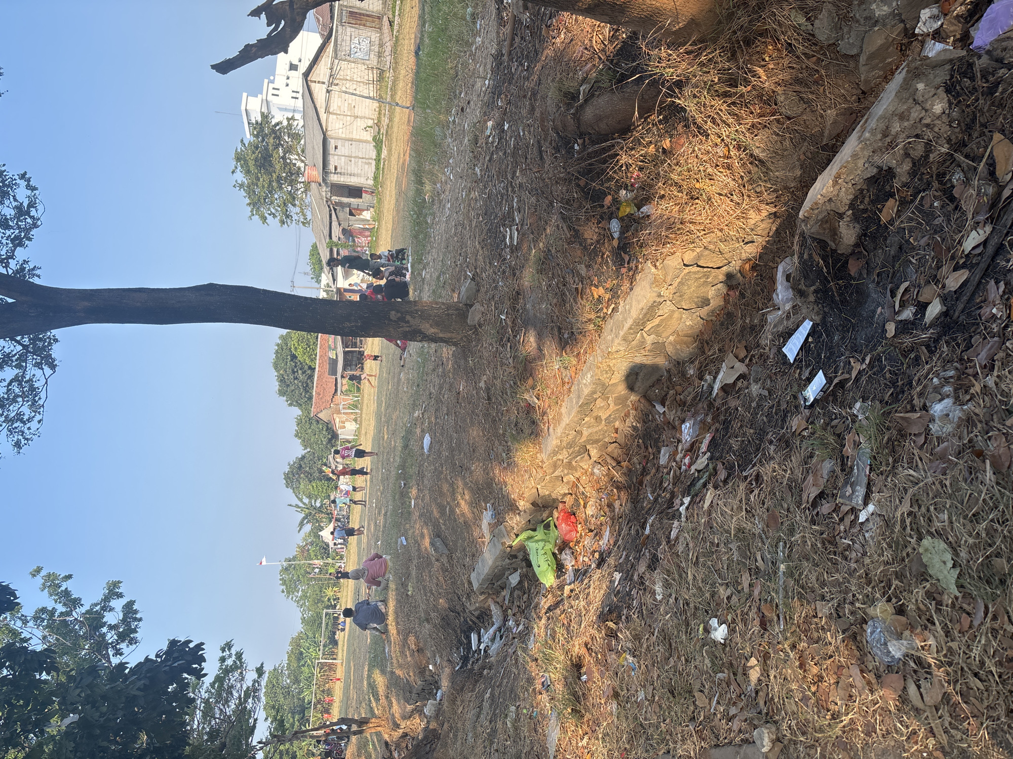The image depicts an outdoor scene in Telagamurni, Indonesia, characterized by a dry, grassy field littered with various types of trash, including plastic bags, wrappers, and debris. A shallow, concrete-lined ditch runs through the foreground, also filled with refuse. In the middle ground, several people, including adults and children, are scattered across the field. Some individuals are sitting, while others appear to be standing or walking. A soccer goal can be observed in the background, suggesting the field may be used for recreation. The environment appears to be a semi-urban or rural area, with modest buildings visible in the distance. The sky is clear and blue. The lighting suggests it is daytime, with the sun casting shadows.