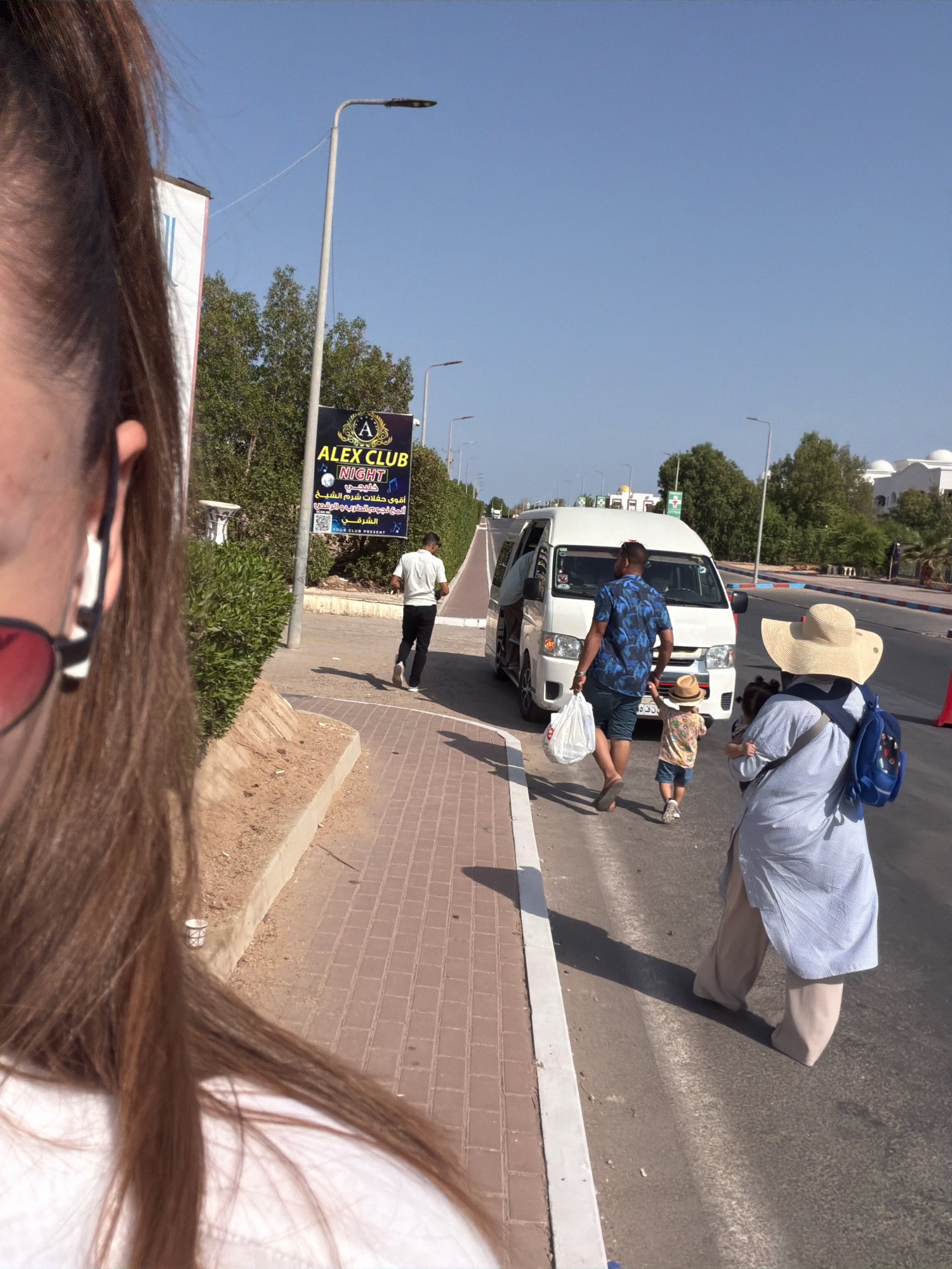 A street scene in Sharm El Sheikh, Egypt, shows a white van stopped on the right side of the road. A man in a blue patterned shirt and shorts is walking away from the van, carrying a white plastic bag. He is followed by a small child wearing a hat and a light-colored t-shirt. To the right of the van, a woman wearing a wide-brimmed hat and a blue and white striped long shirt is holding a small child and carrying a blue backpack. A sidewalk with red brick paving is visible on the left side of the road, with a hedge and a light pole. A sign advertising "ALEX CLUB NIGHT" is mounted on the light pole. The sky is clear and blue, and the sun is shining, casting shadows on the ground. The foreground is partially occupied by the shoulder and hair of a person taking the photo, and a glimpse of sunglasses.