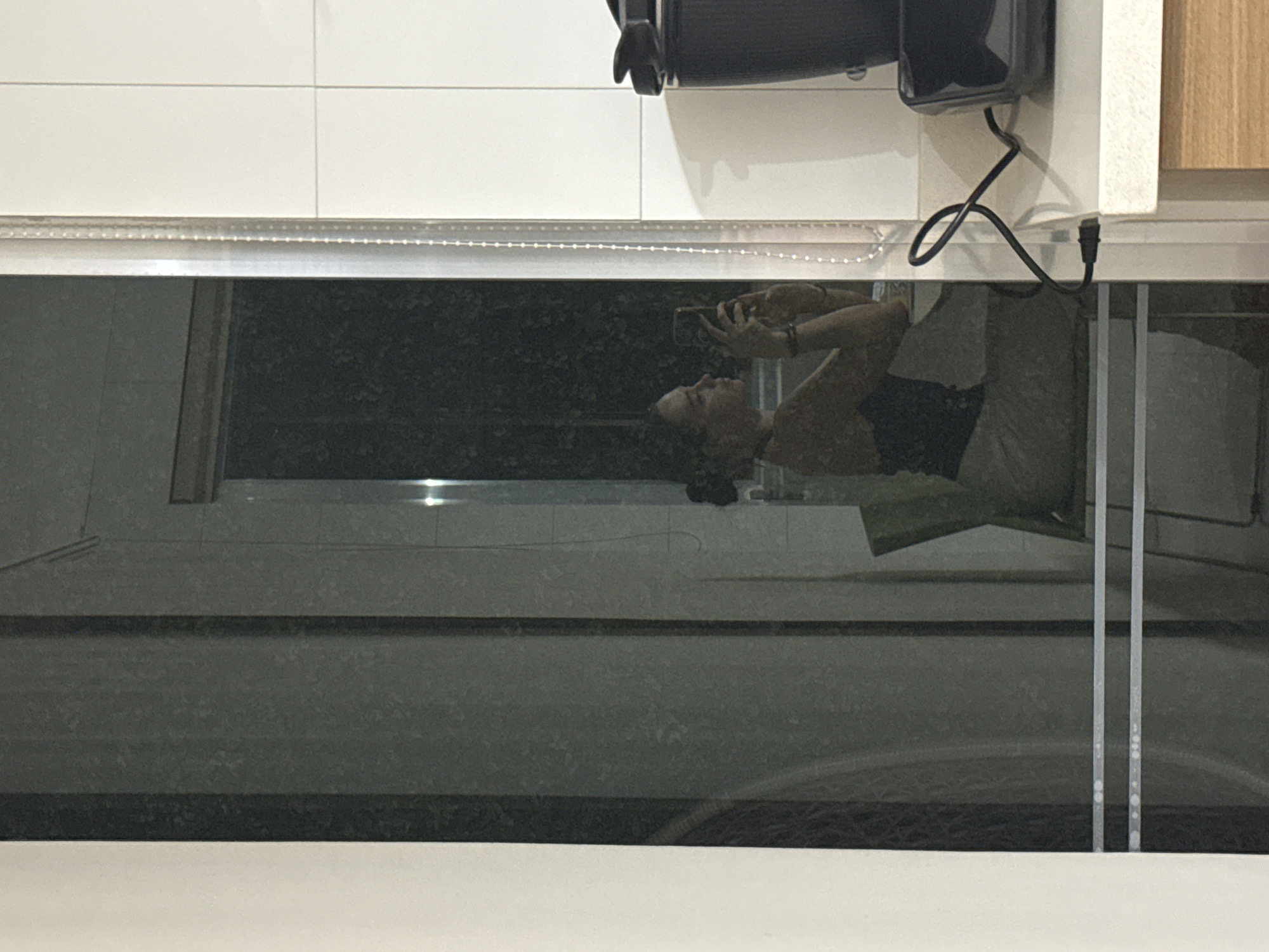 A person is reflected in a shiny black surface, likely a countertop or table. The reflection shows the person holding a mobile phone. The surface also reflects a room. Above the reflective surface, there are white tiles, a black object which appears to be a coffee maker and a wooden cabinet. The location in the city of San Bernardino Tlaxcalancingo, Mexico cannot be confirmed.