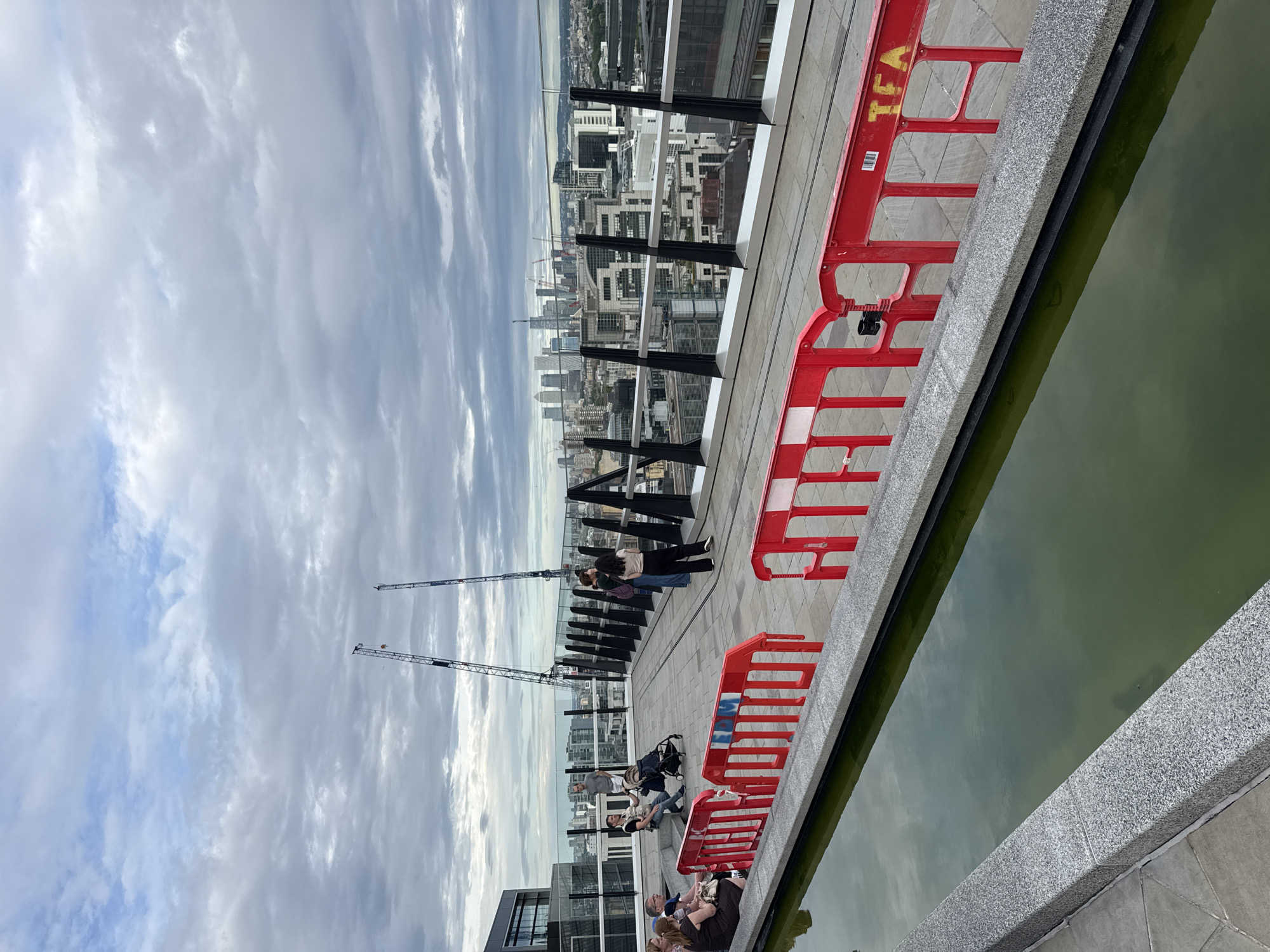 This media file depicts an outdoor scene on a cloudy day, likely in London, United Kingdom. The viewpoint is from an elevated position, possibly a rooftop or balcony, overlooking a cityscape. In the foreground, a body of murky green water runs along a granite-lined edge, bordered by red construction barriers with white markings.
Further into the scene, a paved area features several people. Some individuals are seated or standing, appearing to be engaged in conversation or simply enjoying the view. Others are closer to a glass barrier that separates the rooftop from the city below. Two construction cranes are visible in the distance, suggesting ongoing development in the urban environment. The sky is filled with dramatic, overcast clouds.
The overall mood is somewhat subdued due to the weather, but the presence of people indicates a public space accessible to visitors. The cityscape in the background includes numerous buildings of varying heights, characteristic of a major metropolitan area. One of the red barriers has some yellow text on it, but it is partially obscured and difficult to read clearly. The lighting suggests it is daytime, but the cloudy conditions prevent strong shadows.