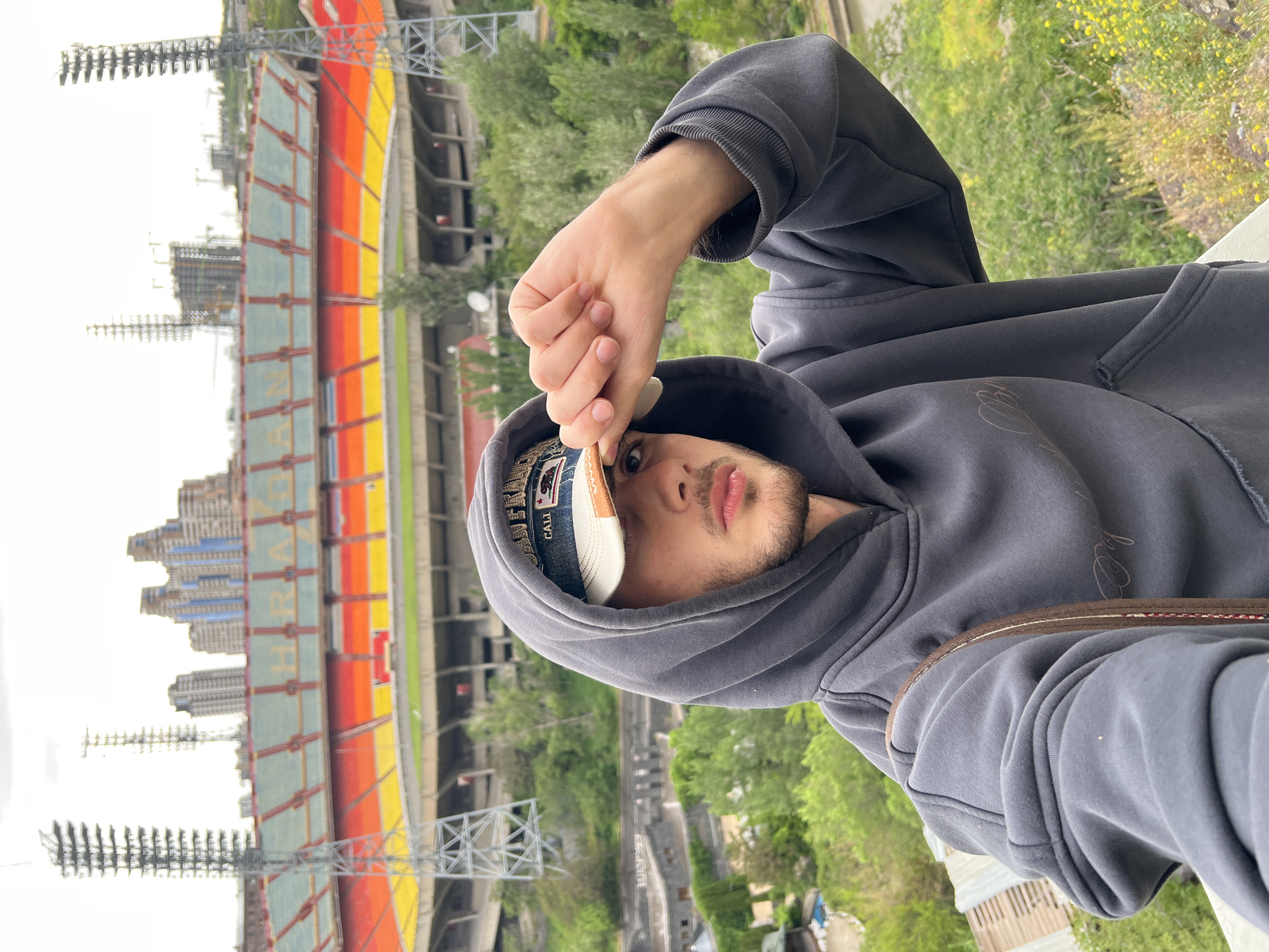 A man wearing a gray hooded sweatshirt and a baseball cap poses for a photo in front of the Hrazdan Stadium in Yerevan, Armenia. He is standing on an elevated platform. The stadium is visible in the background, with its distinct vertical stripes of red, orange, and yellow. In the distance, buildings can be seen on the left side of the stadium. Trees and greenery are visible behind the stadium. The man's right hand is near the brim of his cap.