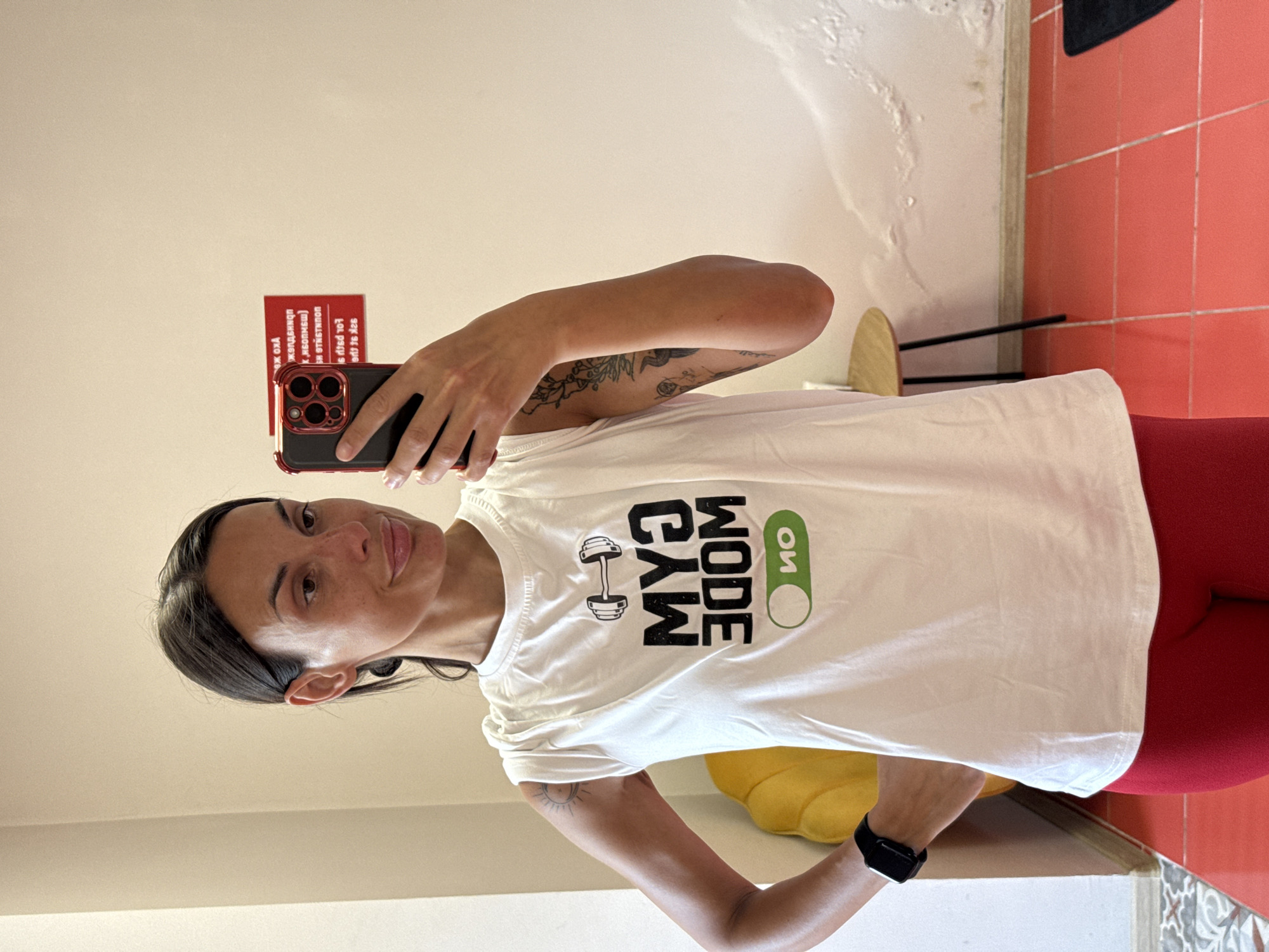 A woman with dark hair pulled back in a ponytail and light skin with freckles is taking a mirror selfie in what appears to be a gym. She is wearing a white sleeveless t-shirt with a graphic that says "GYM MODE" in bold black letters, with a dumbbell icon above and a green "ON" toggle switch below. She is also wearing red leggings and a black smartwatch on her left wrist. Her right hand is holding a phone with a red case, and her left hand is on her hip. In the background, there is a yellow shell-shaped chair, a small round table with black legs, and a wall with peeling paint. A red sign with white text is visible on the wall behind her. The floor is tiled in a reddish-orange color.