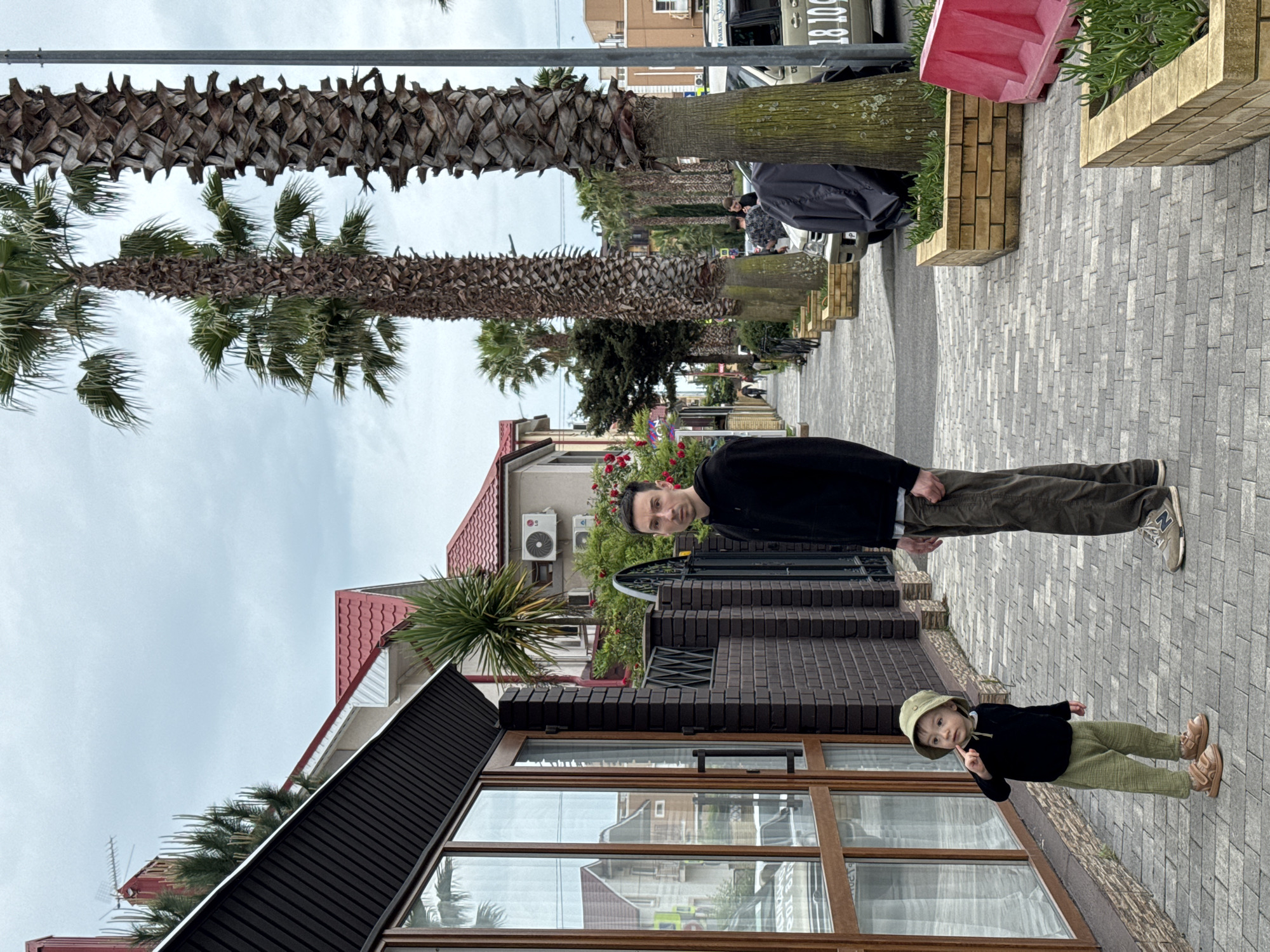 The image shows a street scene in Sirius, Russia. A man and a child stand on a grey brick sidewalk next to a building with large windows and a dark brown roof. A row of palm trees lines the street. Other buildings, including houses with red roofs and air conditioning units, are visible in the background. The sky is overcast.