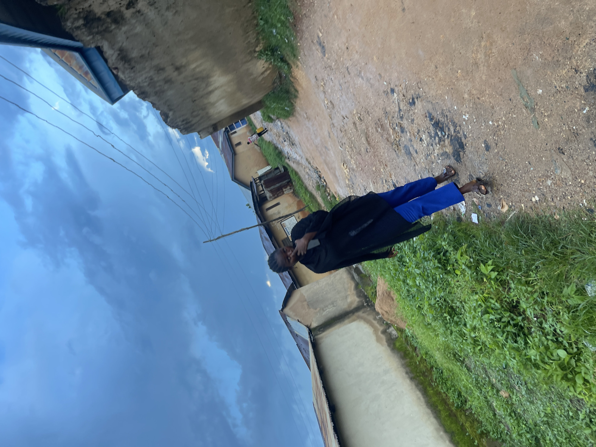 An overcast, cloudy sky dominates the upper portion of this image, casting a diffused light over a dirt road in Rafin Par, Nigeria. The road, appearing somewhat muddy and uneven, winds through a residential area with low-lying buildings on either side. On the left, a pale wall runs alongside the road, bordered by a dense patch of green vegetation. On the right, a rougher, textured wall rises, with another building visible further down the road.
In the foreground, a person dressed in a black garment and blue pants walks away from the camera, their head turned slightly. They appear to be touching their face. Further down the dirt road, another individual is visible, carrying a yellow object. The scene suggests a typical day in a Nigerian neighborhood, with the cloudy sky hinting at the possibility of rain.