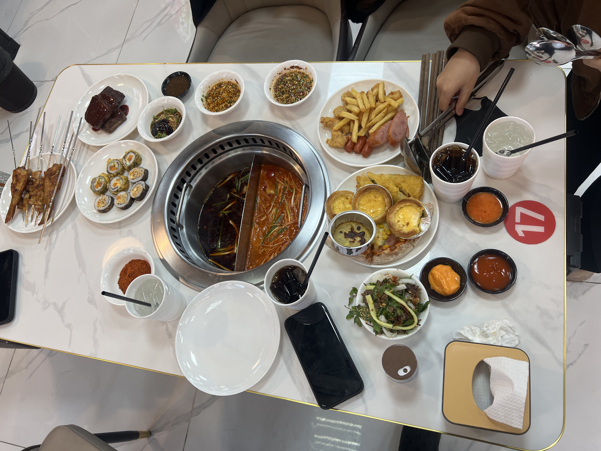 A marble-patterned table with a gold trim is covered with an array of food items in a dining setting. At the center of the table is a split hot pot with two different broths. Surrounding the hot pot, various dishes are arranged including skewers, sushi rolls, dark-colored meat chunks, fries, sausages, egg tarts, and salads. Several small bowls containing sauces and condiments are also scattered around the table. Beverages are present in white cups with straws, and a black smartphone is placed on the table. A tissue box is located on the table's edge. An individual's hand is seen holding chopsticks. The scene likely depicts a hot pot meal at a restaurant. Although there is no information on location in the images, according to the question instructions, it is located in the city of Đà Nẵng, Vietnam.