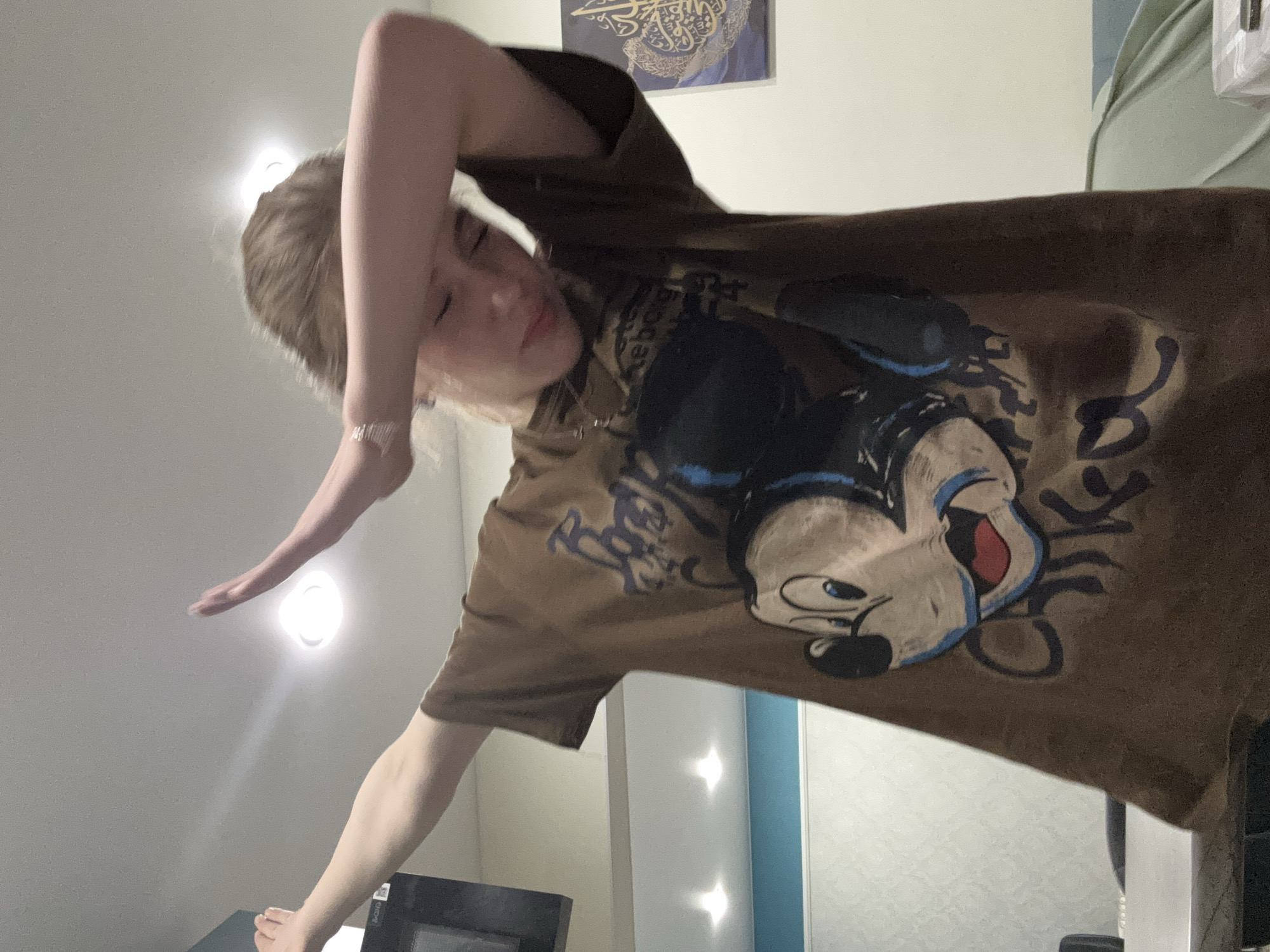 The image shows a young Caucasian female indoors, performing a "dab" pose. She has blonde hair and is wearing a brown t-shirt with a graphic of Mickey Mouse on it. Her right arm is extended upwards, and her left arm is bent across her forehead. She is looking down with her eyes closed and a slight smile on her face. The background appears to be a room with a white ceiling, a light fixture, and a wall with some artwork. There is no indication of the city of Sinyavino, Russia, in the image or its context.