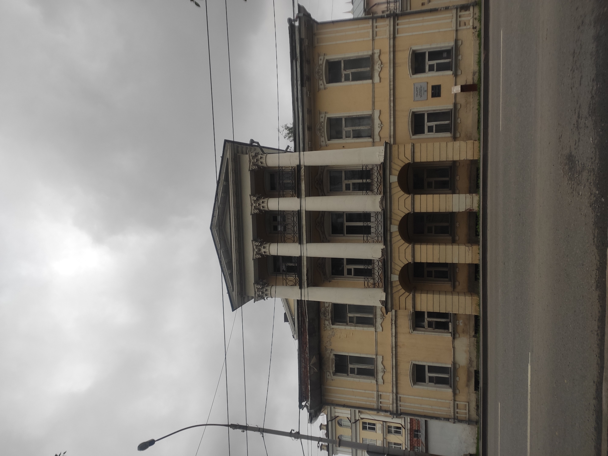 The image shows a neoclassical building with a portico supported by four large columns. The building is painted yellow and has multiple windows. A street light is visible on the left side of the image, and power lines crisscross the sky. The sky is overcast and grey, indicating a cloudy day. The building is situated in front of a road, and there is a patch of grass between the building and the road. In the background, another building with a similar architectural style can be seen. The building appears to be an old public institution. Based on the architectural style and the presence of a plaque on the facade, this building is identified as the Yekaterinburg City Library (formerly the Yekaterinburg City Duma), located at Lenin Avenue, 49, Yekaterinburg, Russia.