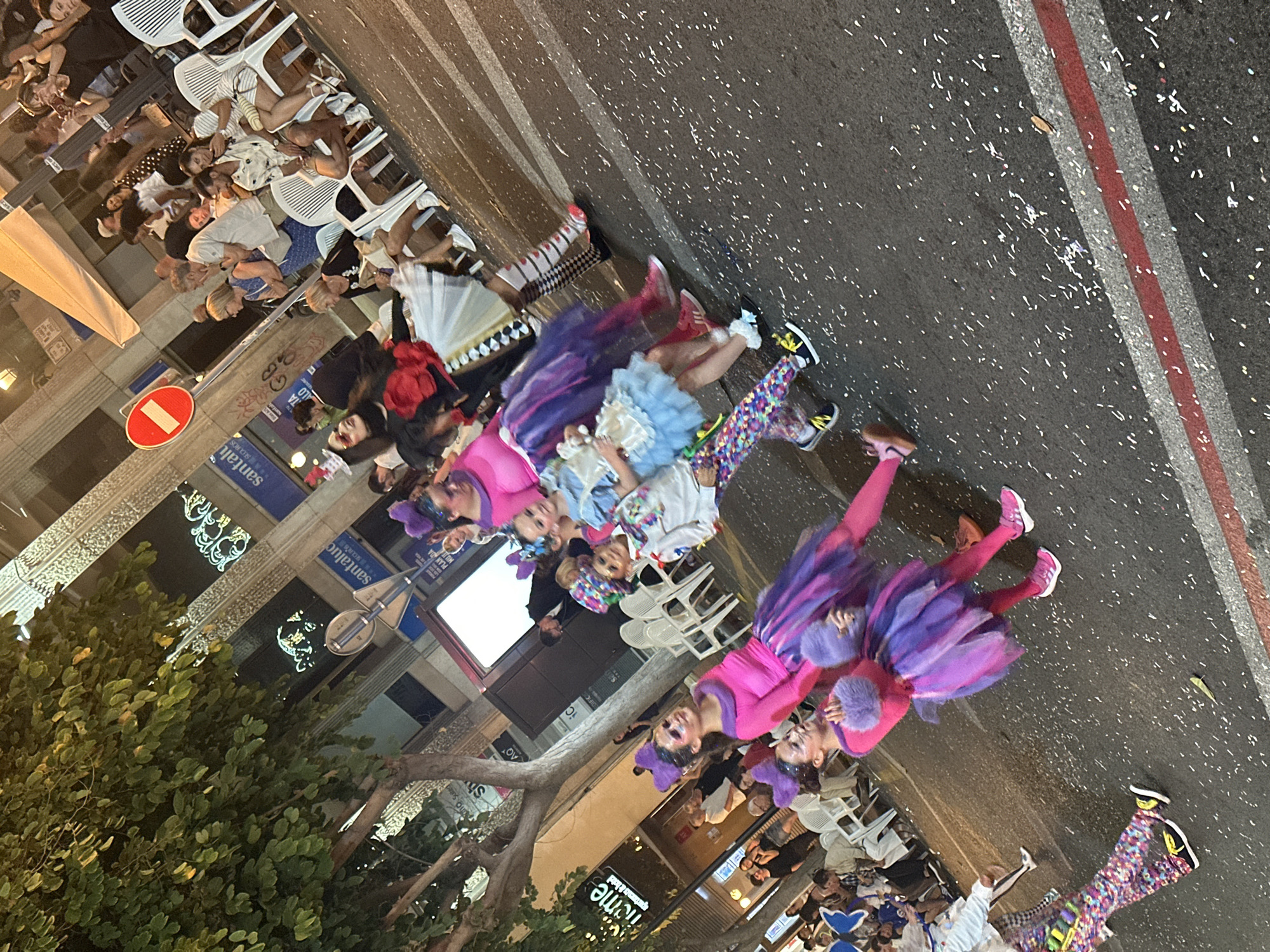 This is a nighttime street scene depicting a parade or festive procession in the city of Alicante/Alacant, Spain.
In the foreground and mid-ground, a group of approximately eight individuals, primarily children and young adults, are walking from left to right along a paved street. They are dressed in elaborate, colorful costumes, consistent with a whimsical or fantasy theme. Key costume elements include:
* Bright pink tops.
* Multi-layered tutu skirts in shades of purple and blue.
* Headpieces resembling animal ears or bows (purple).
* Face paint with white, pink, and purple accents.
* Furry arm details (purple).
* Patterned leggings, some featuring checkerboard designs, hearts, or multi-colored geometric shapes.
* Various athletic shoes, predominantly pink, yellow, or black.
* One adult performer on the right wears a white ruffled skirt, black and white checkered stockings with red heart accents, a dark top, and a large red floral embellishment. They also wear a small, decorative top hat.
* The street itself is dark asphalt, heavily littered with white and multi-colored confetti, indicating an ongoing celebration. Red and grey painted lines mark the edge of the road in the lower right.
On the right side of the image, separated from the performers by a curb, numerous spectators are seated on white plastic chairs, arranged in rows. These individuals, mostly adults, are casually dressed and are observing the procession. One woman in the mid-right is holding a smartphone, seemingly recording or photographing the event. Another woman further down the row has her left lower leg in a white cast, resting it on an adjacent chair.
In the background, the street is flanked by multi-story buildings. Signs for "Santalucía," an insurance company, and partially visible text like "hcmc" and "ato Su" are present. The buildings feature ornate, illuminated decorative elements. A red circular "No Entry" traffic sign is prominently displayed above the spectators on the right. A large tree with dense green foliage occupies the upper left quadrant of the image, partially obscuring the buildings. Artificial lighting from streetlights and building decorations illuminates the scene, casting some shadows.