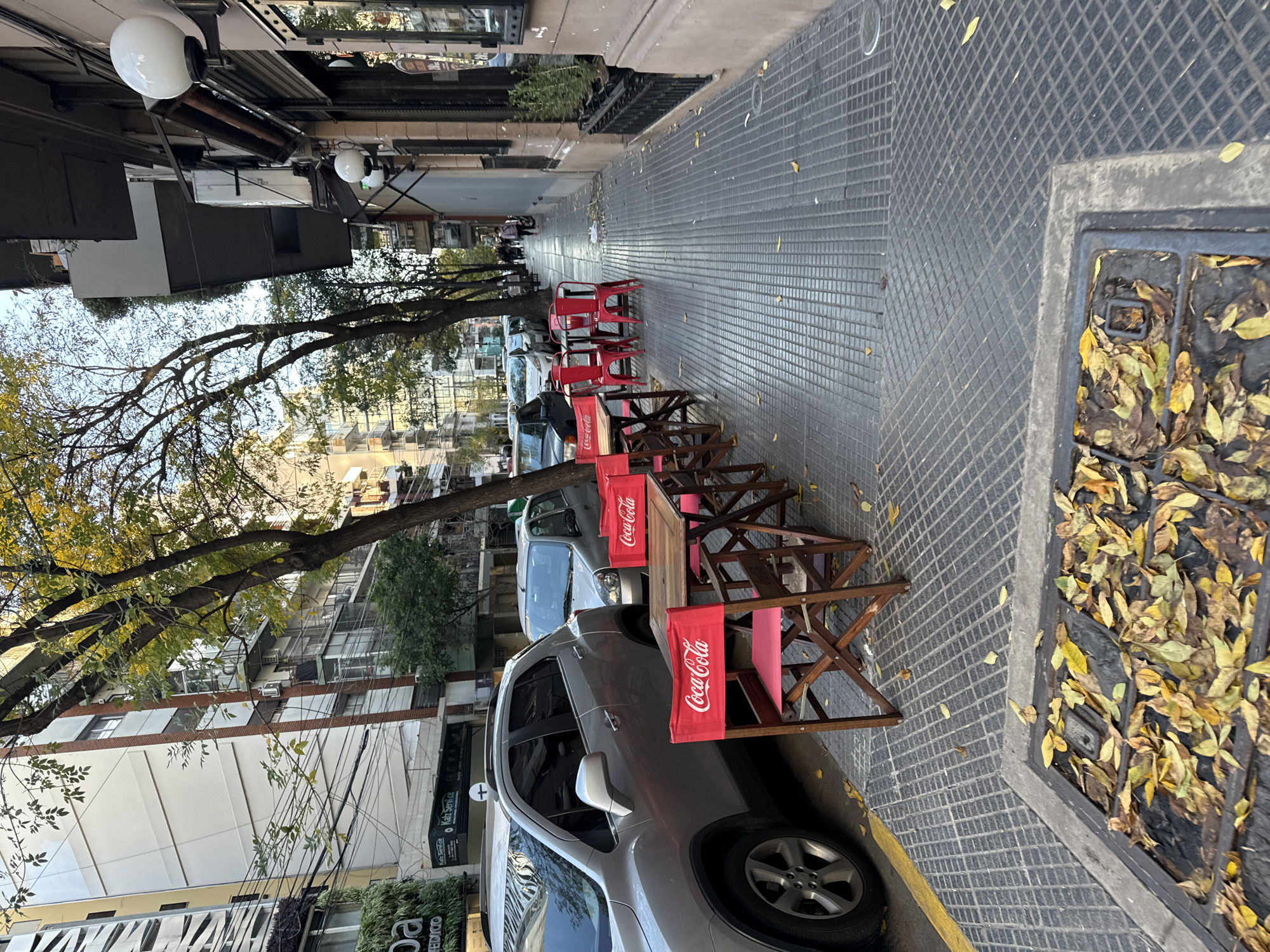 The image captures a street scene in Buenos Aires, Argentina, during autumn. A line of outdoor tables and chairs belonging to a restaurant or café extends along the sidewalk, each chair adorned with a red Coca-Cola branded cloth. Cars are parked along the curb next to the tables, and trees line the street, their branches displaying autumn foliage. The sidewalk is paved with gray, grid-patterned tiles, and a drain filled with fallen leaves is visible. Buildings and other establishments are partially visible on the opposite side of the street, including one with the words "Kalz Service" and "Katz Service" on it, offering some context regarding local businesses. The overall impression is that of a typical Buenos Aires street with outdoor dining options and urban activity.
