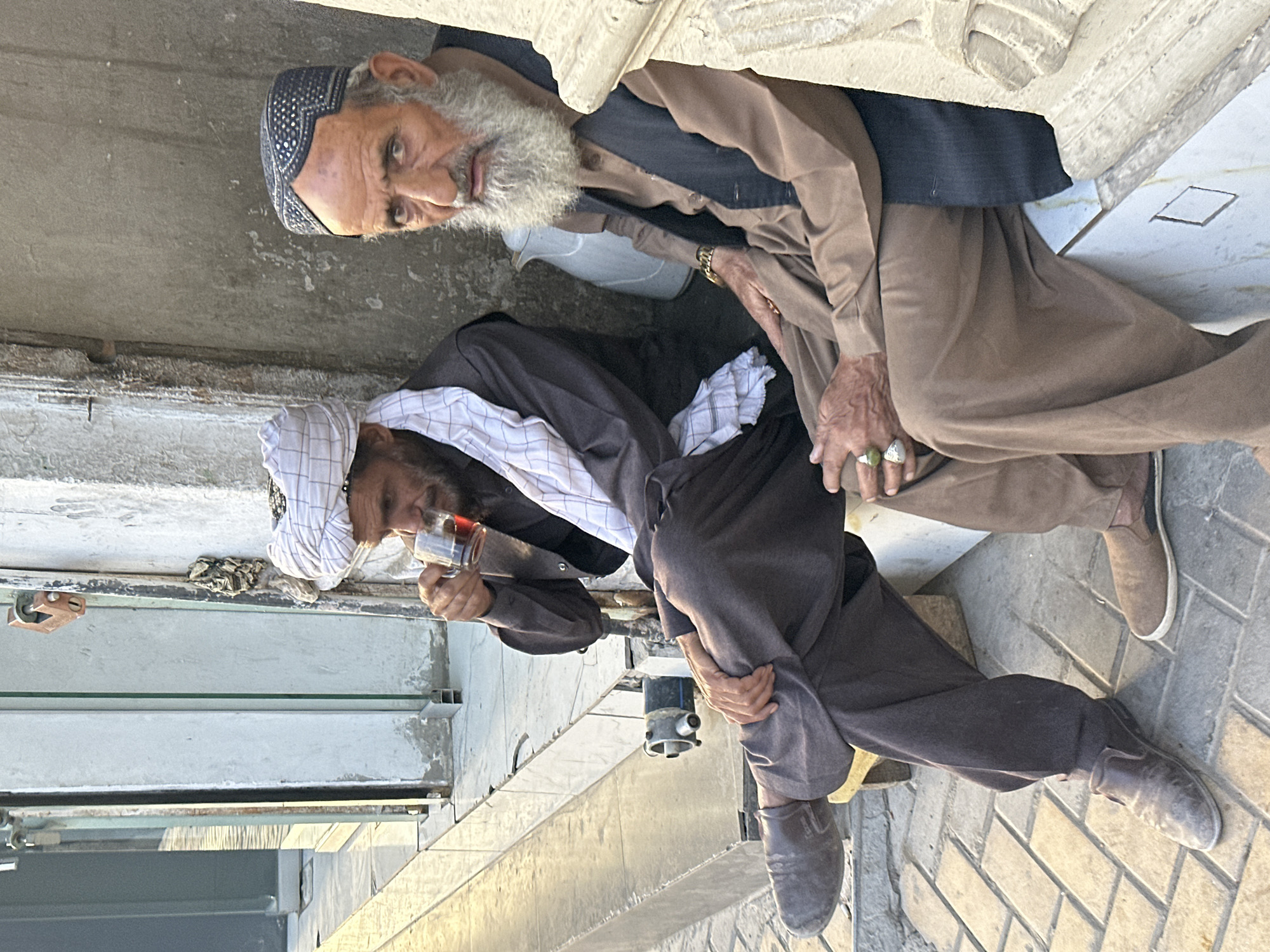 The image shows two older men sitting outside a building in Quetta City Tehsil, Pakistan. One man is wearing a dark cap and a brown coat and vest, and he is looking at the camera. The other man is wearing a white turban and a dark coat, and he is drinking something from a glass. They are sitting on the ground near a wall with tiles.
