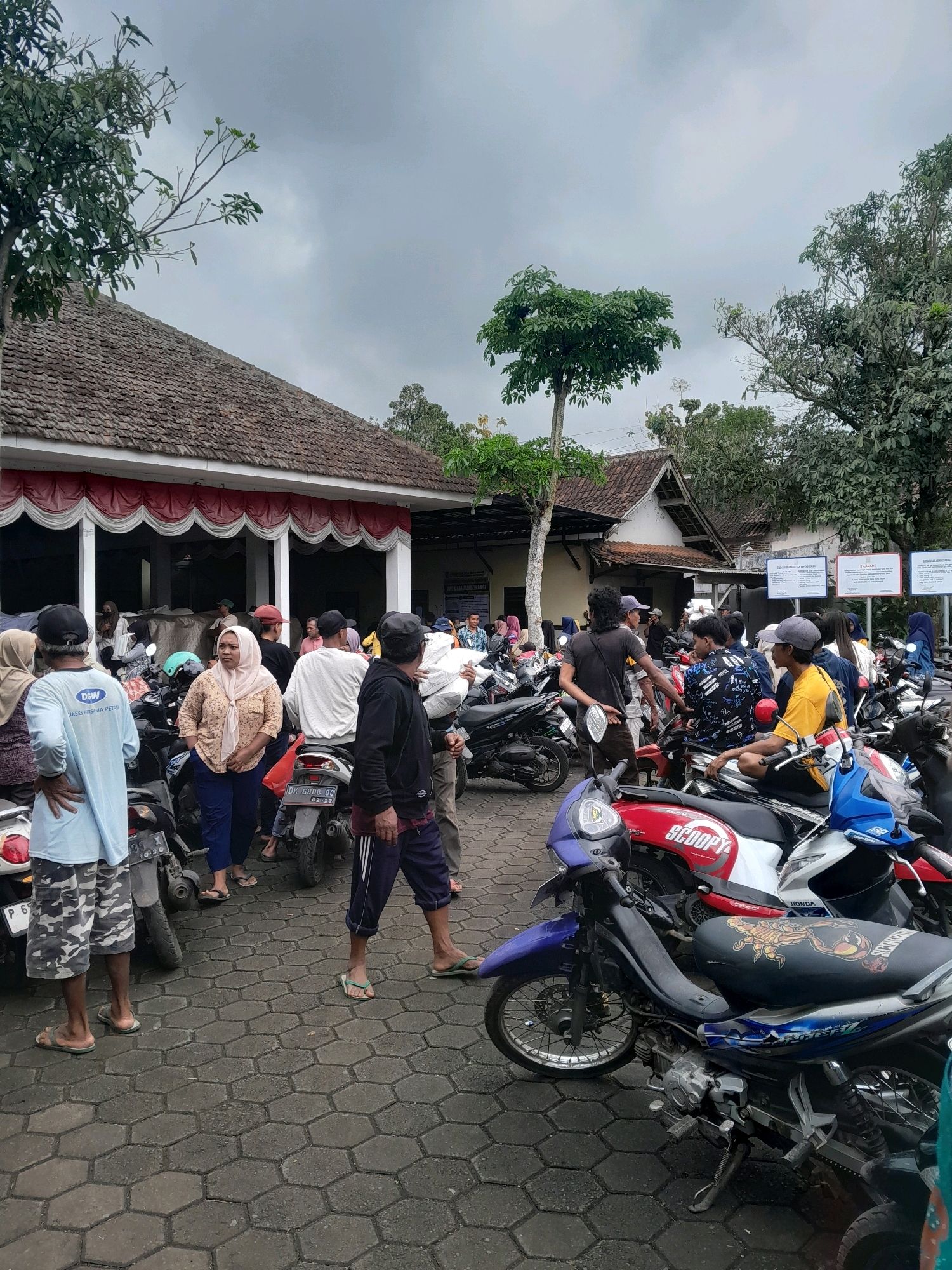 The image depicts an outdoor scene, likely in Jambewangi, Indonesia, characterized by a collection of parked motorcycles and a group of people gathered in front of a building with a tiled roof and a red and white awning. The ground is paved with hexagonal interlocking stones. The sky is overcast and appears to be a cloudy day.
In the foreground, several motorcycles are prominently displayed. The closest is a blue and purple scooter with a black seat featuring a design. To its right is a red and white "SCOOPY" branded scooter. Further back, more motorcycles are visible, including a black one with a license plate reading "DK 6008 OO".
A diverse group of individuals are present, most of whom are standing or sitting on or near the motorcycles. They are dressed in casual attire, with some wearing headscarves or caps. Notable individuals include a person in a light blue t-shirt with camouflage shorts, another in a light pink headscarf and dark pants, and a person in a black hoodie with dark pants. Some are engaged in conversation, while others appear to be waiting. One individual in the foreground is holding a white bag.
The building in the background has a traditional tiled roof and appears to be a public facility or gathering place. Signs with text are visible on the wall of the building, suggesting it is an official location. Lush green trees and foliage are present around the building and in the background, indicating a semi-rural or suburban environment. The overall impression is of a busy, everyday scene in a community setting.