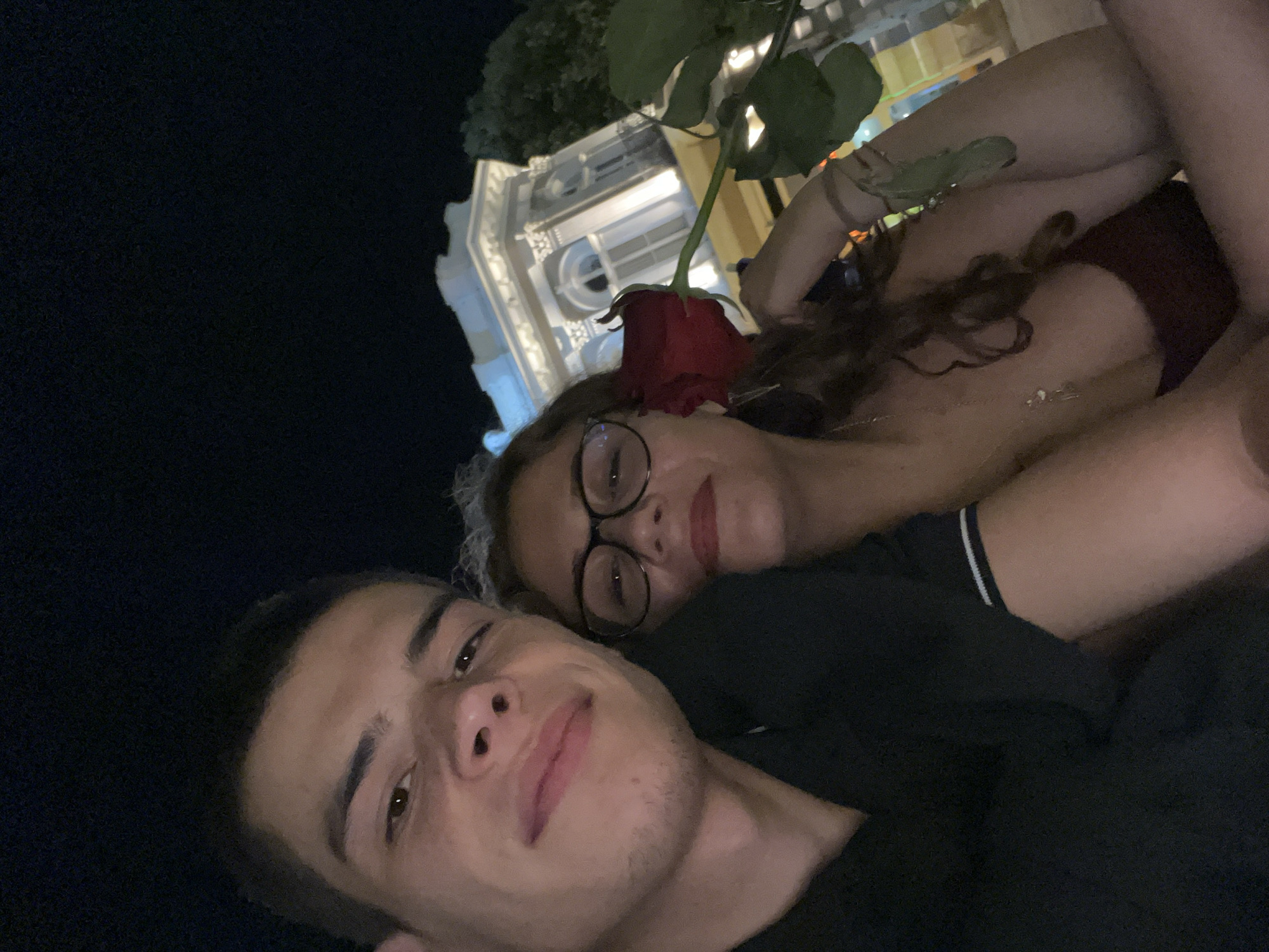 A close-up, low-angle selfie shows a young man and a young woman. The man is in the foreground, smiling slightly at the camera. He has short dark hair and light skin. The woman is behind him, leaning her head on his shoulder, also looking at the camera. She wears glasses and a red rose tucked behind her ear. A building in the background is partially visible, illuminated with white light. The scene appears to be at night in Plovdiv, Bulgaria, likely near the Central Square, based on the architecture of the visible building in the background.