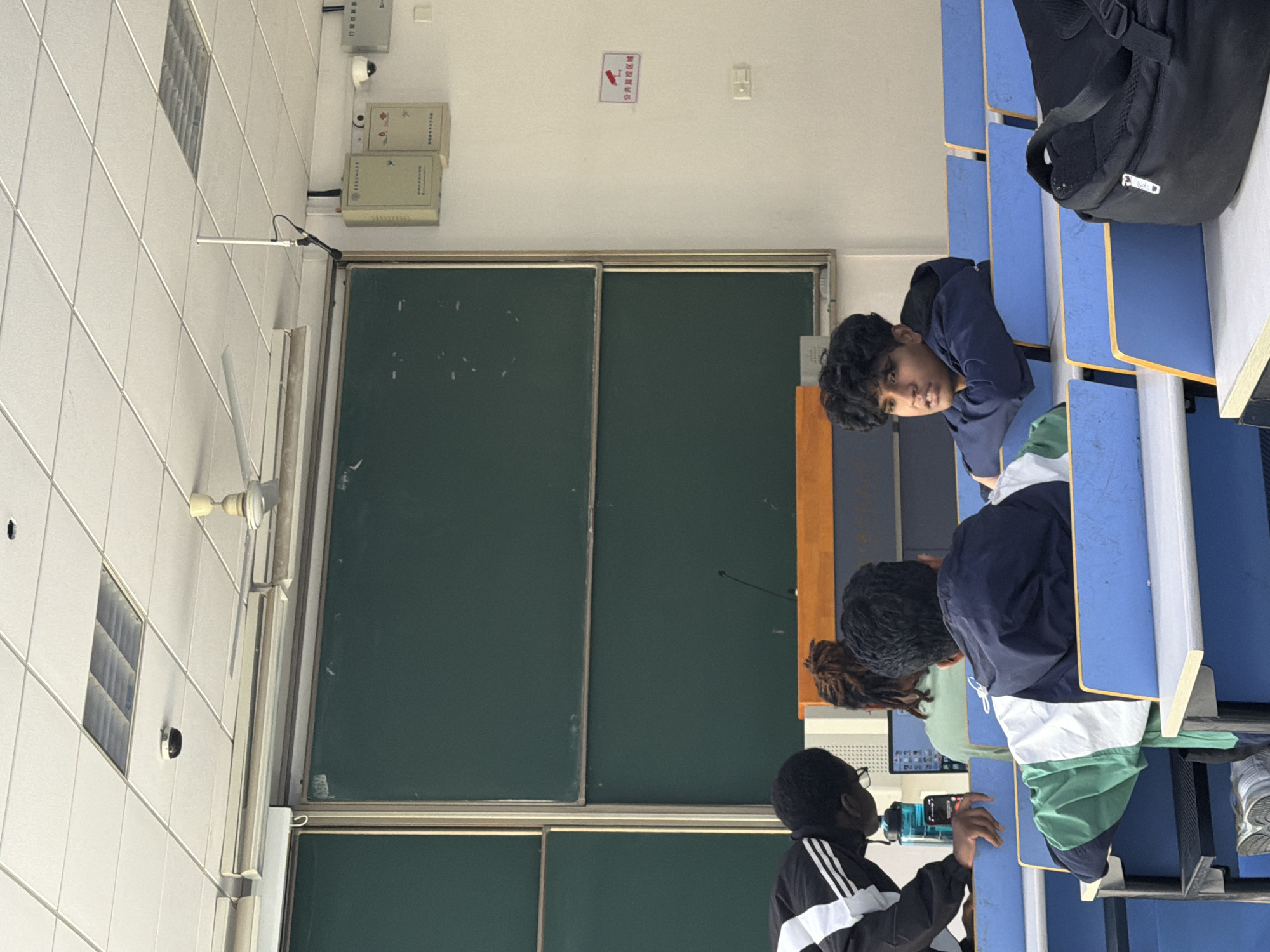 The image shows an interior scene, likely a classroom, with students present. The walls are tiled, and there's a chalkboard with what appears to be two sections. Overhead, there are various fixtures, including a light, a fan, and a white box, possibly electrical in nature. A sign with Chinese writing is mounted near the chalkboard. The students are seated in a row of blue desks, and at least one is looking towards the viewer. The environment suggests an educational setting. I cannot determine the location within the Jiangning District of China.