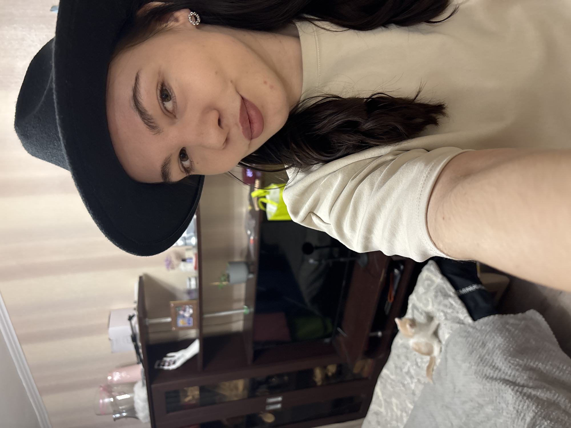 A selfie shows a young woman indoors in what appears to be a living room or bedroom, located in Perm, Russia. The image is taken at an angle, with the subject's head tilted. She has dark hair and brown eyes, looking towards the camera with a slight, neutral smile. She is wearing a black wide-brimmed hat and a light-colored, possibly beige or cream, long-sleeved top. A silver-colored, intricate stud earring is visible in her right ear.
The background features a dark brown wooden entertainment unit or cabinet with shelves. On the cabinet, a small framed picture, a clear glass container, and other indistinct items are visible. A television screen, appearing to be off, is centered within the unit. The wall behind the furniture has a subtle striped pattern in light tones. In the bottom right corner of the frame, a light-colored, possibly white or cream, small cat or kitten is lying on a textured grey blanket or throw, resting on a sofa or bed. The lighting suggests an indoor setting, likely during the day or with ambient artificial light. No visible text is discernible in the image.