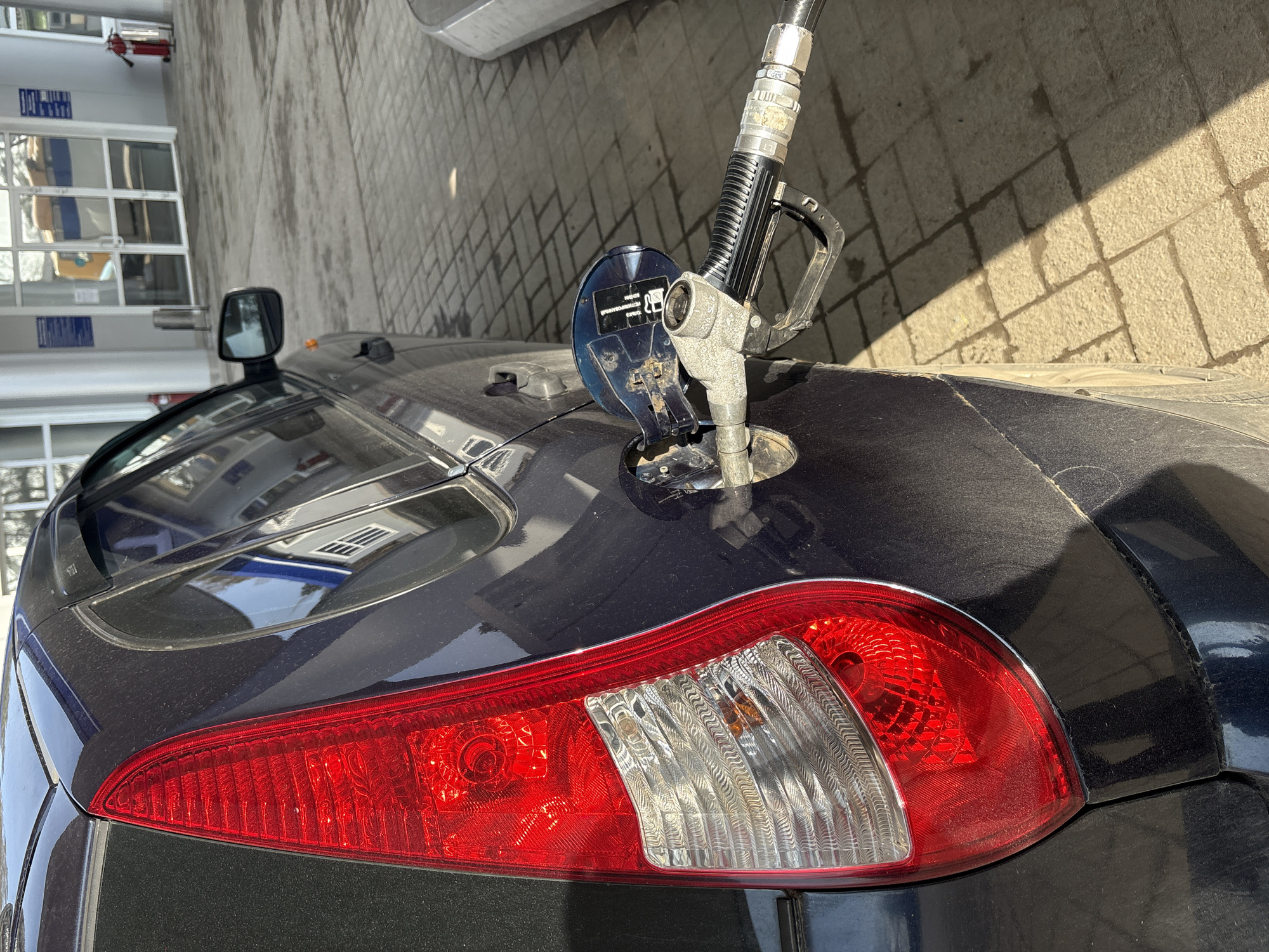 A dark-colored car is being refueled at a gas station. The gas pump nozzle is inserted into the car's fuel tank. The car's rear tail light is visible, with red and white sections. The setting appears to be in Chaikovsky, Russia based on the metadata. The background shows a building with windows, likely part of the gas station. The ground is paved with light-colored tiles.