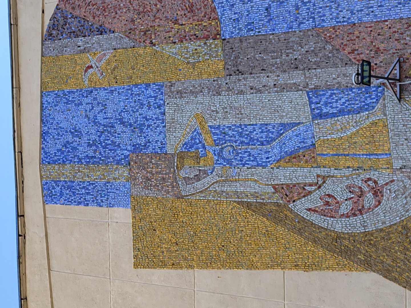 The image shows a mosaic mural on a building in Chaikovsky, Russia. The mural depicts an abstract composition of geometric shapes and figures. Dominant colors include shades of blue, yellow, red, and brown. The patterns suggest stylized representations of objects and people. A small black object, possibly a camera or sensor, is mounted on the mural's surface. The building's facade is made of light-colored concrete or stone.