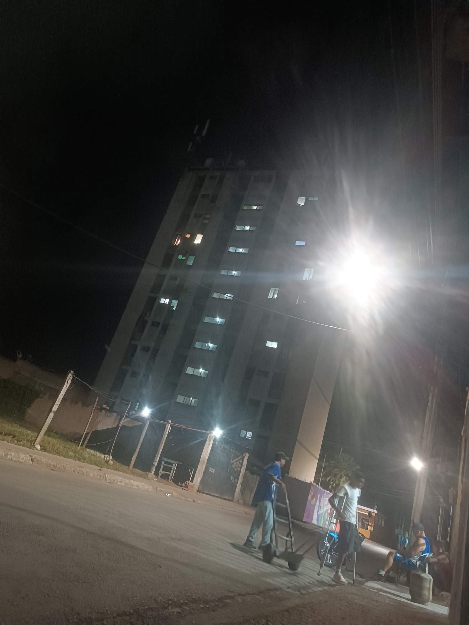 The image depicts an outdoor nighttime scene on a street in Porlamar, Venezuela. A tall, light-colored multi-story building, likely residential, occupies the left-center background, with numerous illuminated windows. The street runs across the foreground, delineated by a curb and an unpaved grassy area to the left, which is bordered by simple wooden or concrete posts. Several artificial light sources, including a prominent streetlight on the right, create significant lens flares across the frame.
Four male individuals are present. On the left, one man wearing a blue shirt and light trousers pushes a two-wheeled handcart or dolly away from the camera. Towards the center-right, another man in a white t-shirt and dark shorts stands supported by crutches, looking towards the right. A bicycle with a distinctive wheel design is positioned nearby. Further to the right, two more men are seated. One, wearing a blue tank top and patterned shorts, sits on a chair or stool next to a visible propane tank. The other individual is partially obscured, seated lower, to his right. The men on the right appear to be resting or socializing. The environment suggests an urban setting with a mix of residential structures and street-level activity.