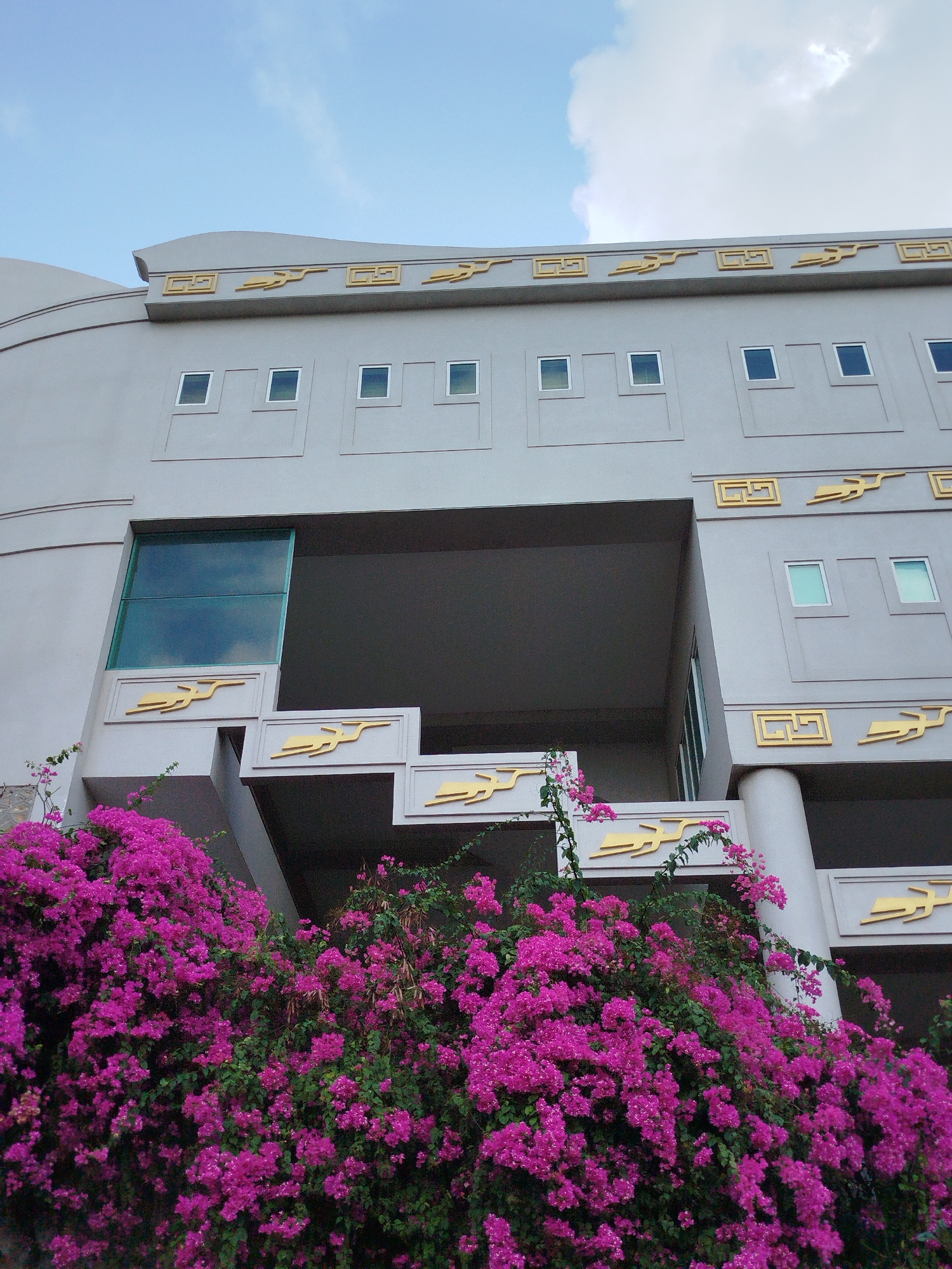 A low-angle shot shows a section of a multi-story, light gray building, likely in Ho Chi Minh City, Vietnam, adorned with decorative gold accents resembling stylized birds and geometric patterns. The building's design features a central open space on the lower levels. A vibrant mass of bright pink bougainvillea flowers cascades over the stepped lower section of the building, partially obscuring the architectural details. The sky above is visible, showing patches of blue with light clouds. Windows are small and square, spaced evenly along the facade. The building appears modern, with simple lines and minimal ornamentation beyond the gold accents.