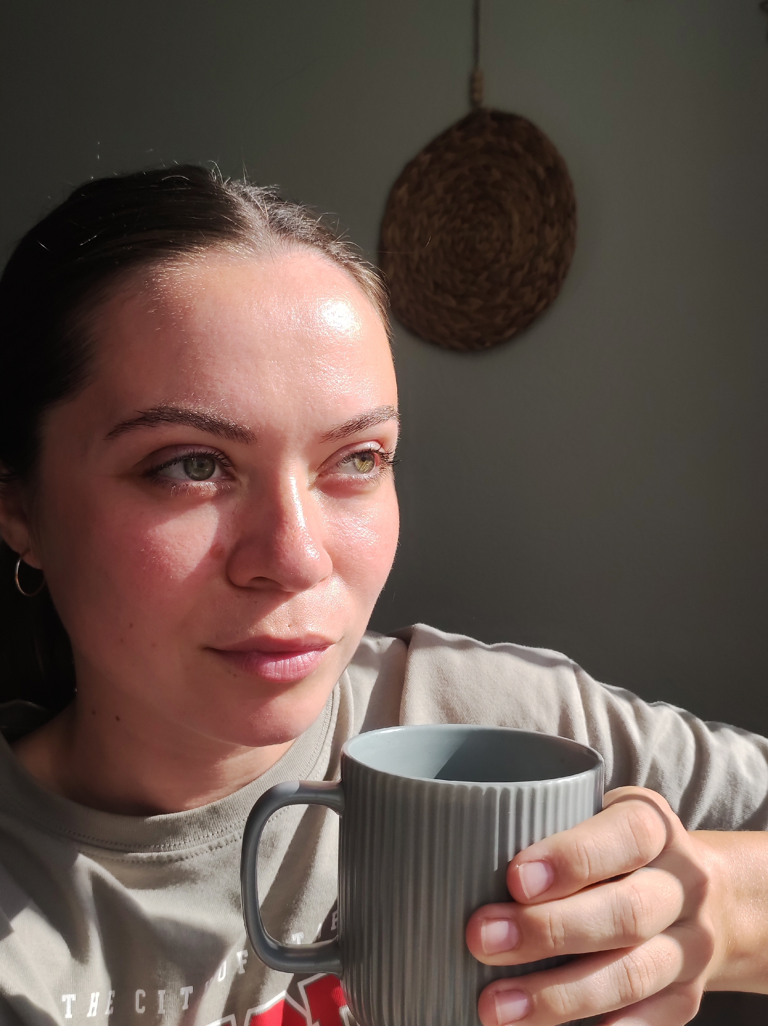 A medium shot portrait shows a young woman with light skin, green eyes, and dark hair pulled back. She is holding a gray mug with a ribbed texture in her right hand. The woman is looking to her right, with her head tilted slightly. Sunlight illuminates her face, creating highlights on her forehead, nose, and cheeks. She is wearing a light-colored t-shirt with text on it that is partially visible. In the background, a woven circular object hangs on the wall. The scene appears to be indoors, with a plain wall and soft lighting. The image does not contain any information that would allow for its location to be identified as the city of Chaikovsky, Russia.