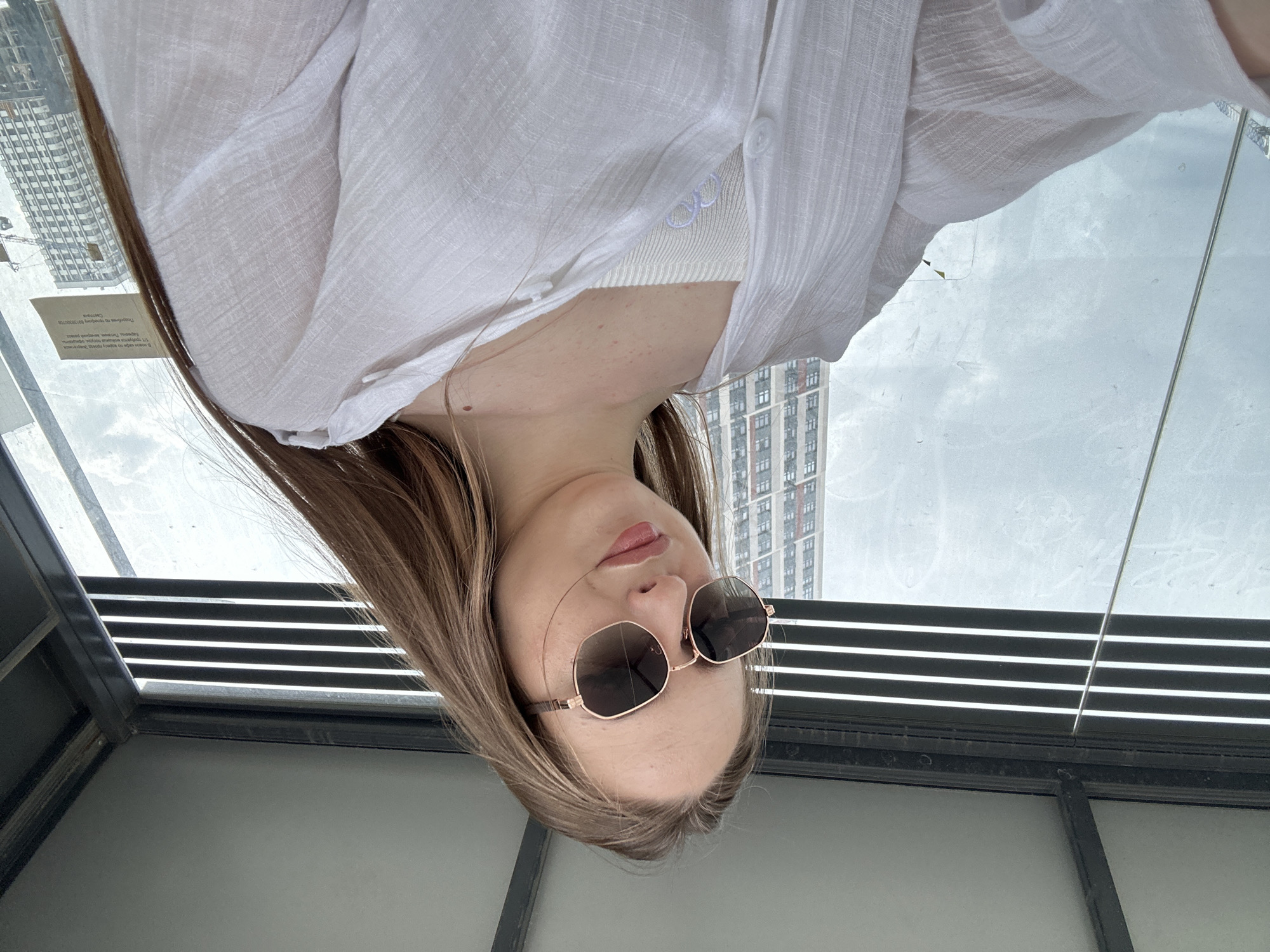 A young woman with long, brown hair and sunglasses is taking a selfie. She is wearing a white shirt and a white tank top underneath. She is indoors, possibly in a bus stop or sheltered area, with a large window behind her showing a cityscape with tall buildings. The lighting is overcast.