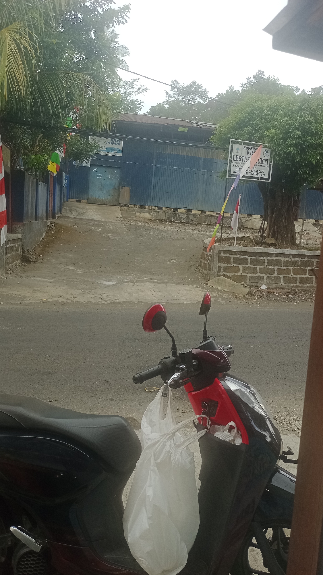 This image depicts an exterior scene in what appears to be a commercial or industrial area, potentially in Cikalong, Indonesia. In the foreground, a red and black motorcycle is parked, with a large white plastic bag hanging from its handlebars. The motorcycle is positioned on a paved road. Beyond the road, there is a dirt or gravel driveway leading up to a building with a blue corrugated metal facade. A white sign with text is visible on the building. Another sign, reading "KOOPERASI LESTARI," is mounted on a brick wall to the right of the driveway. In the background, lush green trees and foliage are present, suggesting a tropical or subtropical environment. The sky is overcast. The image composition suggests it was taken from another vehicle or a standing position close to the motorcycle.