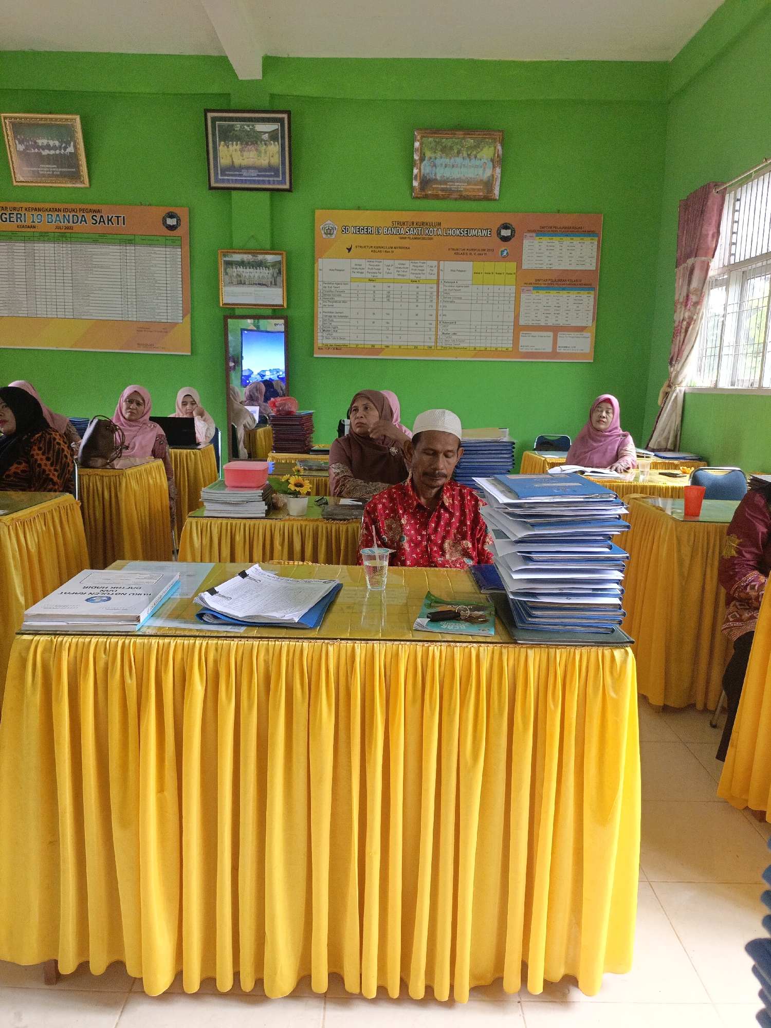 The image shows an indoor scene that appears to be a classroom or meeting room in Lhokseumawe, Indonesia. The walls are painted a vibrant green. On the walls are several framed photographs and posters, including a list of "DAFTAR URUT KEPANGKATAN (DUK) PEGAWAI NEGERI SIPIL KOTA LHOKSEUMAWE" and a "STRUKTUR KURIKULUM".
In the foreground, there are several tables covered with bright yellow tablecloths. On these tables are stacks of papers, folders, books, and a few plastic cups. A man wearing a red and white patterned shirt and a white cap is seated at one of the tables, looking down with his eyes closed. Several women, mostly wearing headscarves in pink and brown, are also seated at tables in the room, some looking at laptops or at the documents in front of them. In the background, there is a window with a patterned curtain. The floor appears to be tiled.