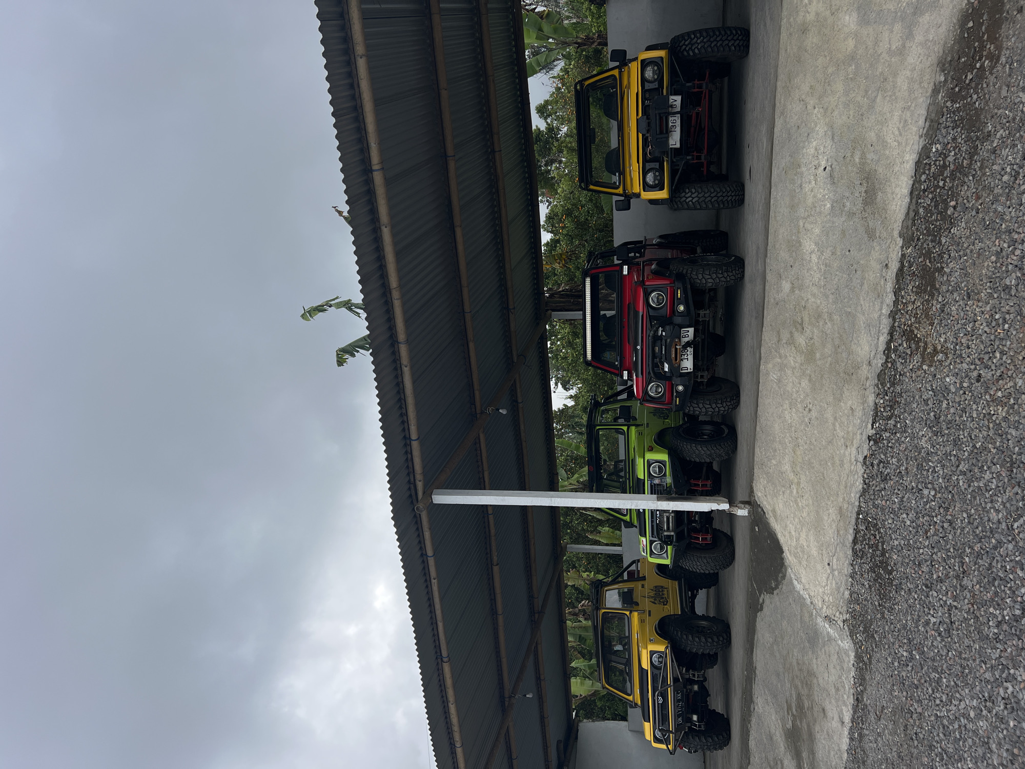 A group of four off-road vehicles are parked under a corrugated metal roof supported by wooden beams and white concrete pillars. The vehicles, from left to right, are yellow, lime green, red, and yellow. All are modified with large tires and off-road bumpers. The ground beneath the vehicles is a paved surface, with a gravel area visible in the foreground. In the background, lush green vegetation, including banana trees, is visible. The sky is overcast with grey clouds. The location is the city of Kintamani, Indonesia.