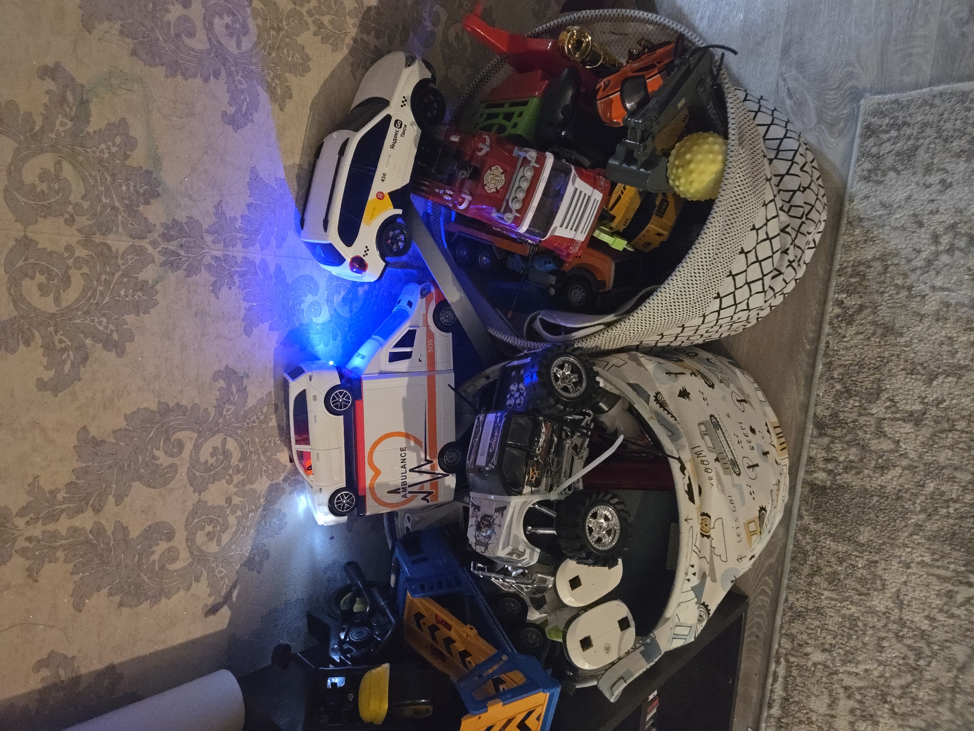 The image depicts a collection of toy vehicles and other objects stored in two fabric baskets, placed on a carpeted floor. The toys are primarily cars, including an ambulance, a white SUV with taxi markings, and a large toy truck. Some of the toys are illuminated by blue light. The background features a textured wallpaper with a decorative pattern. The overall scene suggests a child's play area. There is no indication of the city of Vladimir, Russia, in the provided image or any details that would allow for its identification as such.