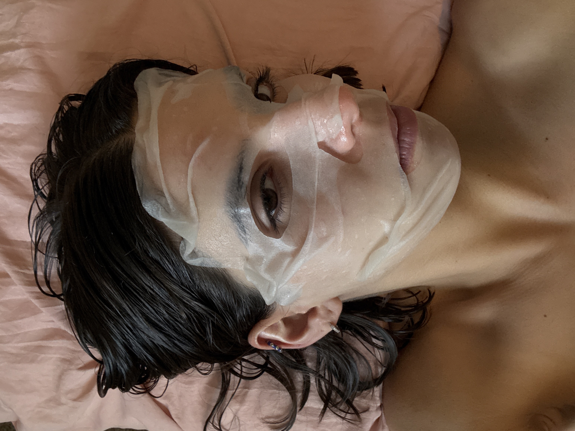 A woman lies on a pink pillow in Southlake, Texas, USA, wearing a white sheet mask. Her wet, dark hair is scattered on the pillow around her head. She has an earring in her left ear, and parts of her upper chest and shoulder are visible. The focus is on her face, which is partially covered by the mask.