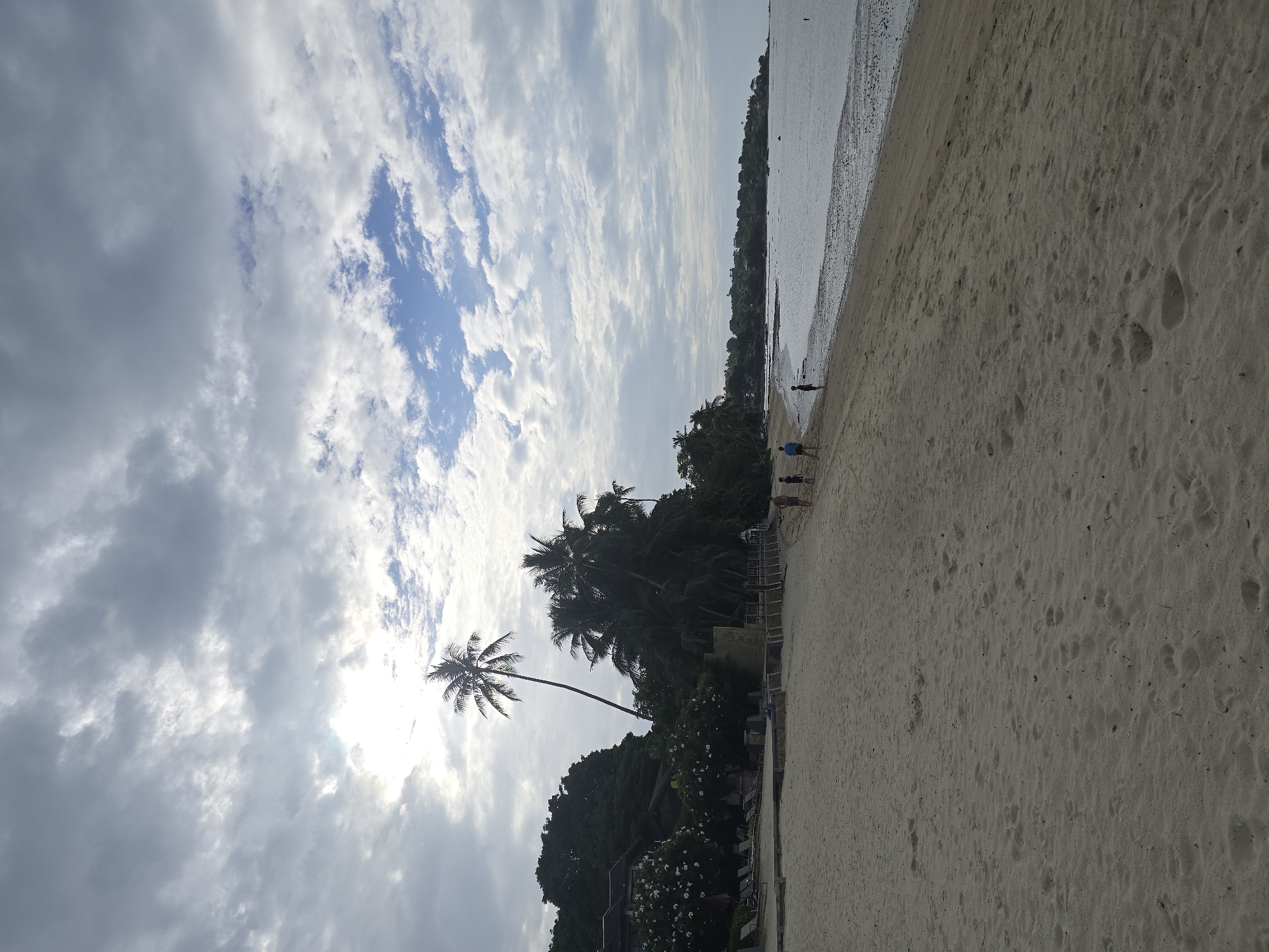 A vertical, low-angle shot captures a serene beach scene under a cloudy sky with patches of blue. The foreground is dominated by a sandy beach with numerous footprints. Further back, a calm body of water meets the shore. A line of palm trees and lush green foliage borders the beach, with some buildings visible amidst the greenery. The sky is filled with a mix of dark and light gray clouds, with the sun peeking through in the upper left corner, casting a bright glow. Several people are walking along the shoreline in the middle ground.