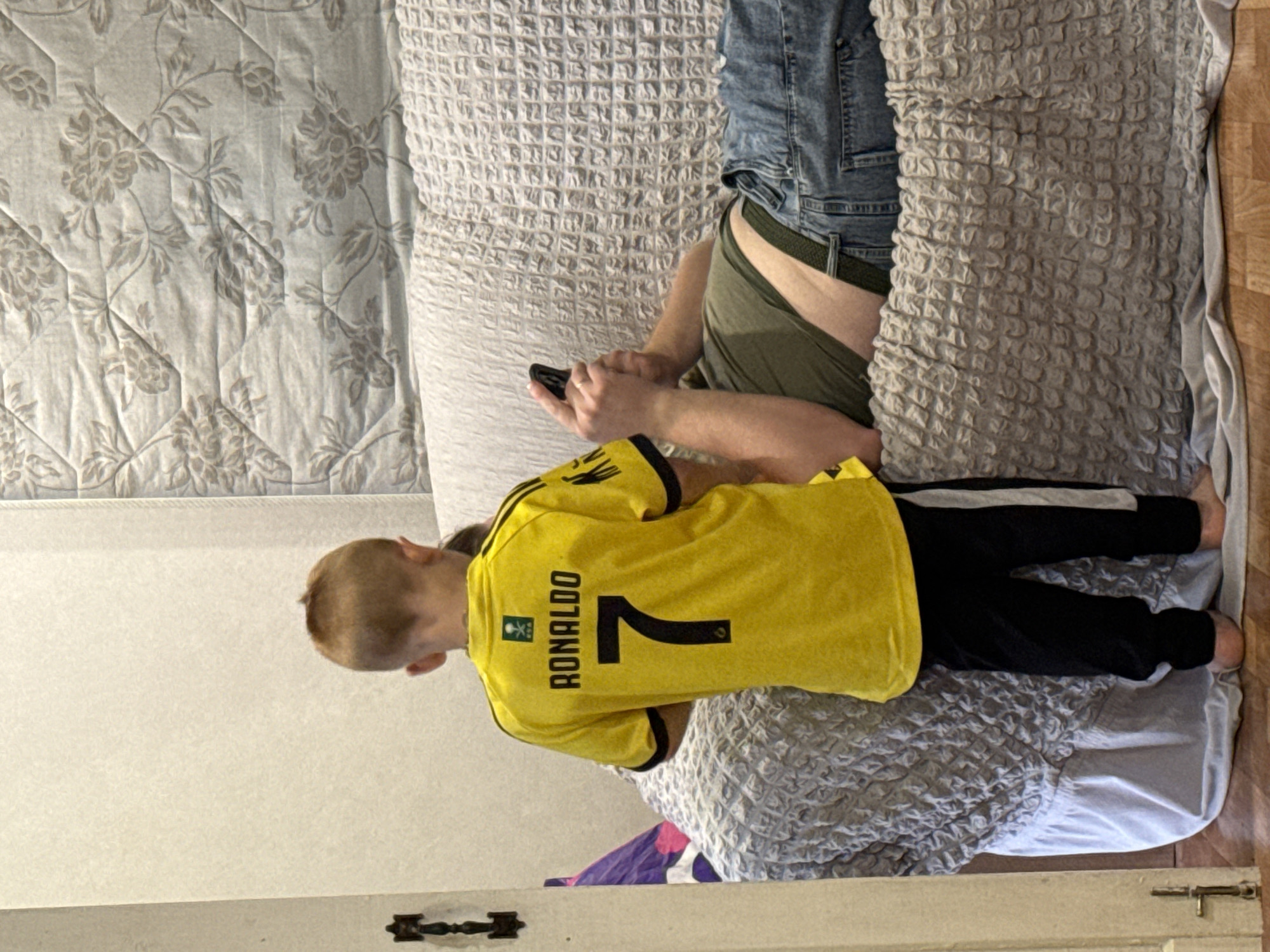 A young boy wearing a yellow Ronaldo jersey and black pants stands facing away from the camera, looking at a person lying on a couch. The person on the couch is wearing a green t-shirt and blue jeans and is holding a smartphone. The couch is covered with a grey textured blanket. The background consists of a wall and a floral-patterned quilt. The scene appears to be indoors, possibly a living room.