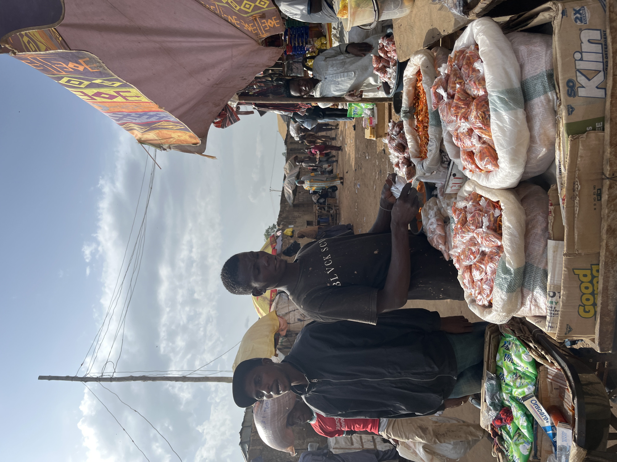 The image shows an outdoor market scene in Rafin Par, Nigeria. Two young men are standing in the foreground behind stalls laden with goods. The man on the left wears a black jacket and a black baseball cap, while the man on the right wears a black t-shirt. In front of them are large sacks filled with small, orange items, likely dried chilies or peppers, some of which are packaged in clear plastic bags. Other sacks and boxes containing similar produce are visible on the stalls. To the left of the young men, another person is partially visible carrying a large sack on their head. In the background, several other people are walking through the market, and various market stalls and structures are present, including awnings and what appear to be buildings made of mud brick. The sky is partly cloudy.
