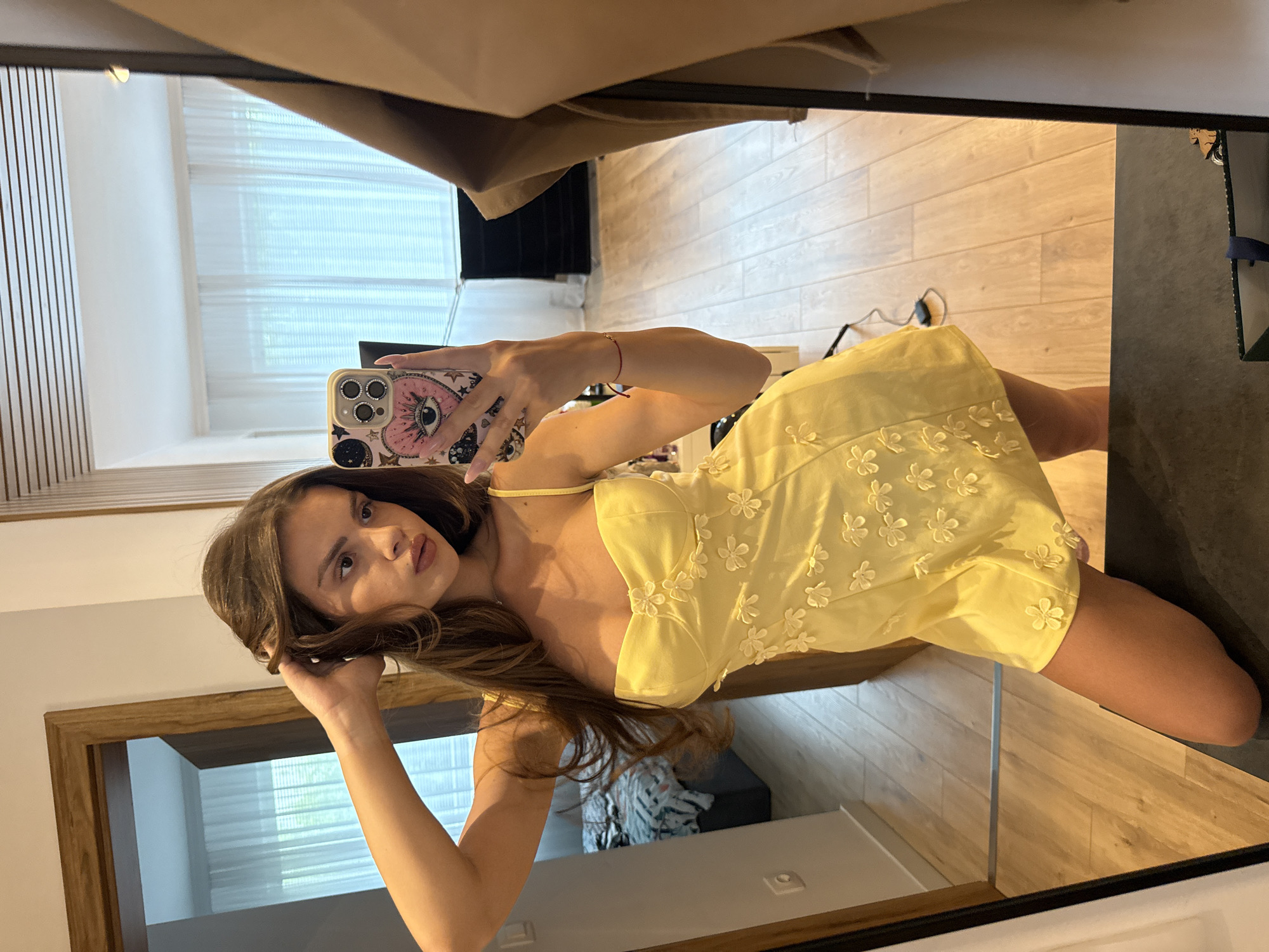 A young woman with long brown hair is taking a mirror selfie in a room. She is wearing a yellow dress with flower embellishments and holding her hair with one hand while holding a phone in the other. The room appears to be a bedroom or dressing area, with a doorway, curtains, and wooden flooring visible in the reflection. There is no indication of the city of Plovdiv, Bulgaria.