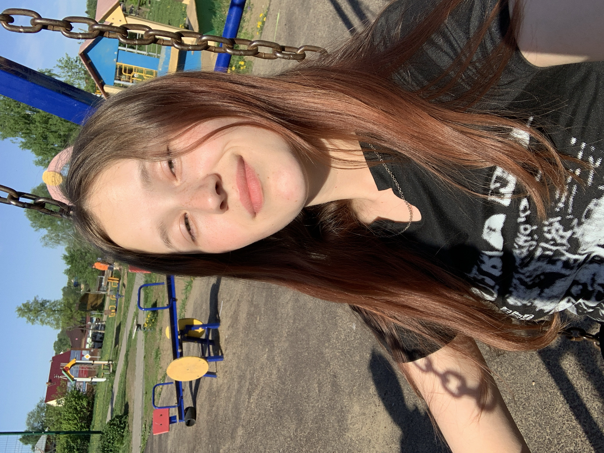 A young woman with long, auburn hair is photographed on a swing set in what appears to be a playground. The swing chains are visible at the top of the frame. She is wearing a black t-shirt with a white graphic design on the front. Her face is slightly turned, and she is smiling softly with her eyes partially closed, suggesting the sun is bright. The playground is populated with various pieces of equipment, including a yellow and blue merry-go-round and a blue swing set structure. In the background are buildings and trees, suggesting a residential area. Given the city of Фоки, Russia, the playground is likely within that vicinity.