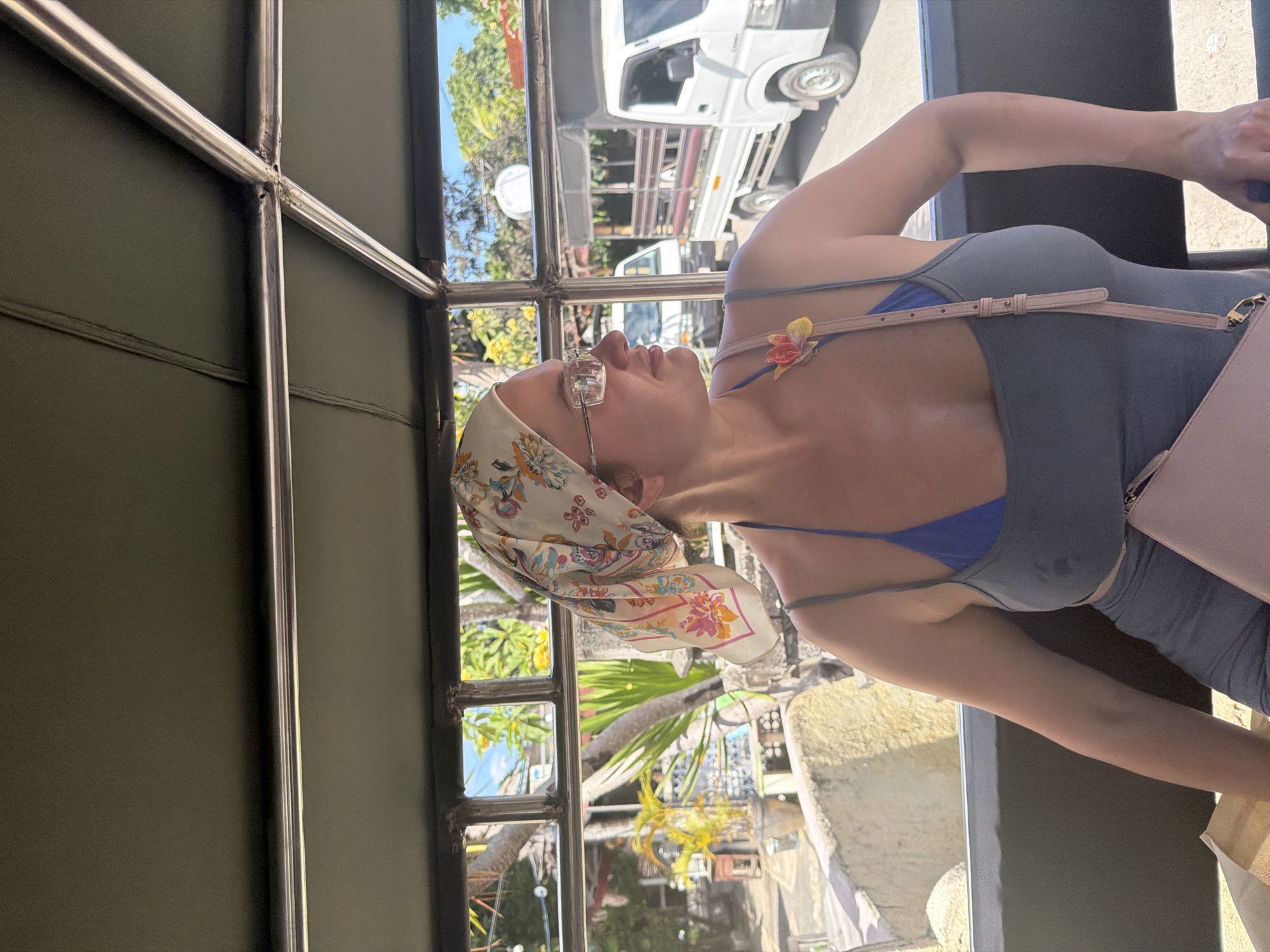 A woman with a floral headscarf and glasses is sitting inside a vehicle, looking to her right. She is wearing a blue bikini top and a grey tank top. A pink crossbody bag is slung over her shoulder. Outside the vehicle, palm trees and other lush vegetation are visible, along with a white truck. The scene appears to be in a tropical location, possibly Nusa Penida, Indonesia, given the presence of tropical flora and the style of the vehicle.