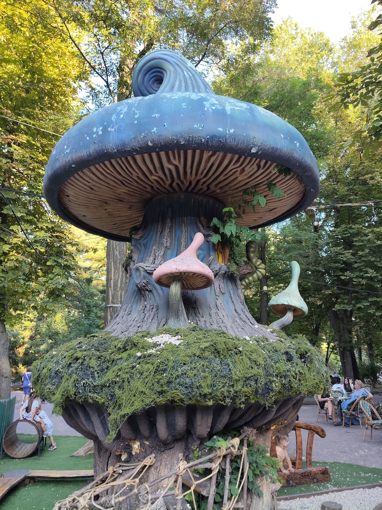 In Chișinău, Moldova, a fantastical, oversized mushroom sculpture dominates the scene in an outdoor park setting. The sculpture features a large, blue mushroom cap with a swirling pattern on top, supported by a thick, textured trunk. Two smaller, distinct mushrooms sprout from the trunk: one a pinkish-red, and the other a pale green, both with curved stems. The base of the sculpture is covered in lush, green moss, with smaller white accents resembling miniature mushrooms or flowers.
Several people are visible in the background. To the left, two children are interacting with a large wooden, barrel-like structure, one sitting inside it and the other perched on the edge. To the right, a group of adults are seated at tables, appearing to be at an outdoor cafe or seating area. They are engaged in conversation and relaxation. The environment is characterized by mature trees with dense green foliage, suggesting a well-established park or garden. String lights are visible overhead, hinting at evening or decorative lighting. The overall atmosphere is one of a whimsical, family-friendly recreational space.