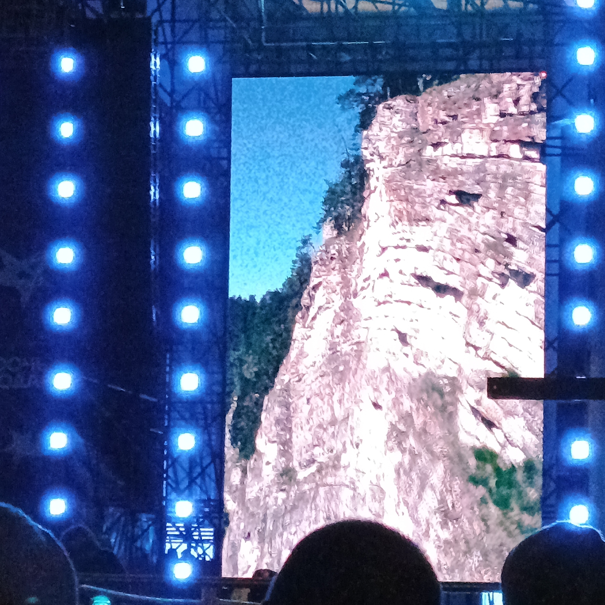 The image depicts a large digital screen displaying a scenic view of a rocky cliff face with greenery and a blue sky. The screen is flanked by vertical rows of bright blue lights. In the foreground, the silhouettes of several people are visible, with their heads and shoulders obscuring the bottom portion of the screen and the stage area. The overall setting appears to be an outdoor event or concert in Perm, Russia, likely at the Perm State Art Gallery or a nearby venue known for hosting large-scale visual presentations.