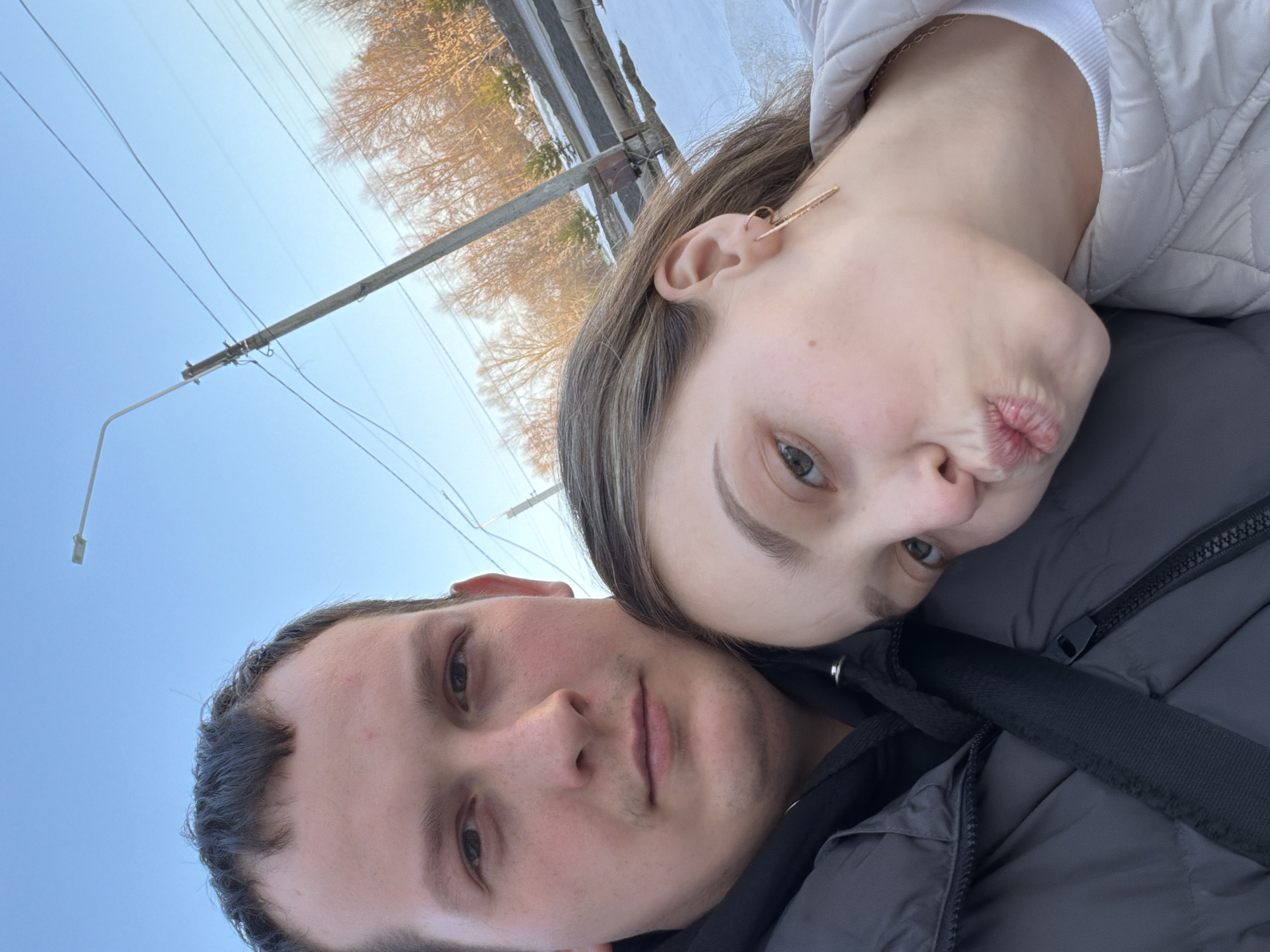 A selfie-style photograph captures a man and a woman outdoors in Chaikovsky, Russia, during what appears to be winter or early spring. The man, positioned on the left and filling the majority of the left frame, has short dark hair, a light complexion with faint facial hair, and wears a dark grey puffer jacket over a black hoodie, looking directly at the camera with a neutral expression. The woman, positioned to his right and slightly lower, leans her head against his shoulder area. She has light brown hair, a light complexion, wears gold-colored dangle earrings, and a light-colored quilted jacket over a white top. Her lips are pursed in a "kissing" pose, and her visible left eye looks slightly upwards. The background features a clear blue sky, a concrete utility pole supporting multiple overhead power lines, and a street light fixture. To the right, a snow-covered ground or embankment is visible, alongside an exposed pipe structure. Deciduous trees with bare branches and some evergreens are present in the distant background, illuminated by sunlight.