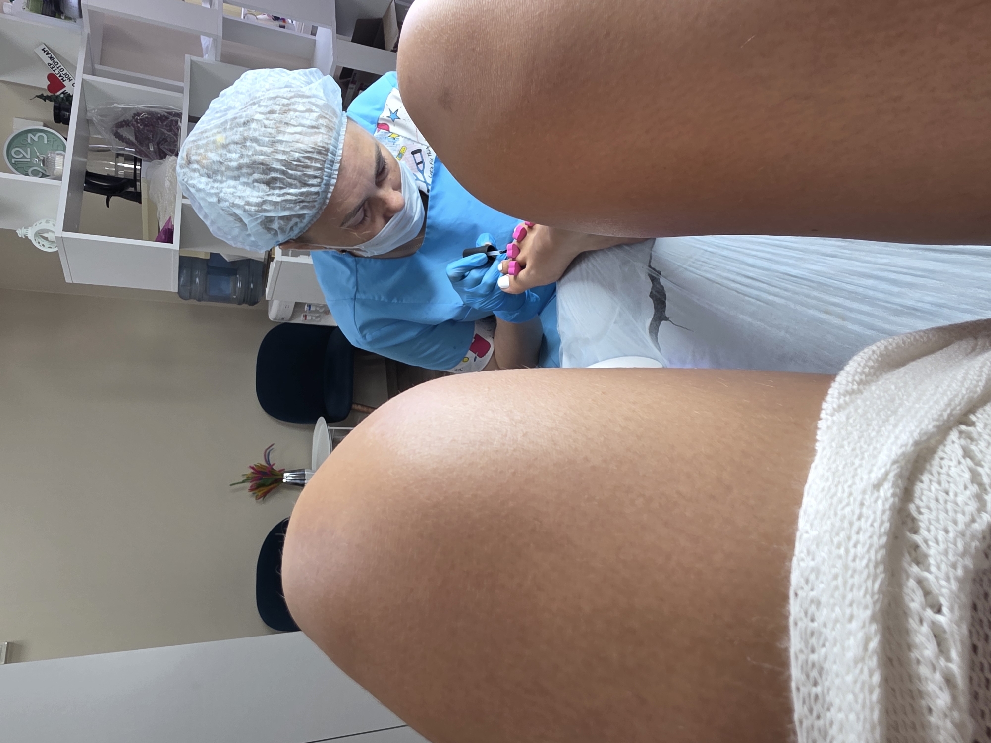 The image shows a close-up view of a pedicure being performed. In the foreground, two legs are visible, with the right leg slightly more in focus. The individual receiving the pedicure is wearing a white knitted garment around their lower thigh. In the mid-ground, a nail technician, wearing a blue uniform, a surgical cap, and a face mask, is carefully painting the toenails of the person on the left. The toenails are partially painted with white and pink polish, with some toe separators in place. The technician is holding a bottle of nail polish and a brush. The background reveals a clean, well-lit salon environment with shelves containing various items, including a water dispenser, decorative objects, and what appears to be a clock. Two dark blue chairs are visible in the background, suggesting a waiting area. The setting is likely a nail salon in Sochi, Russia, given the implied context.