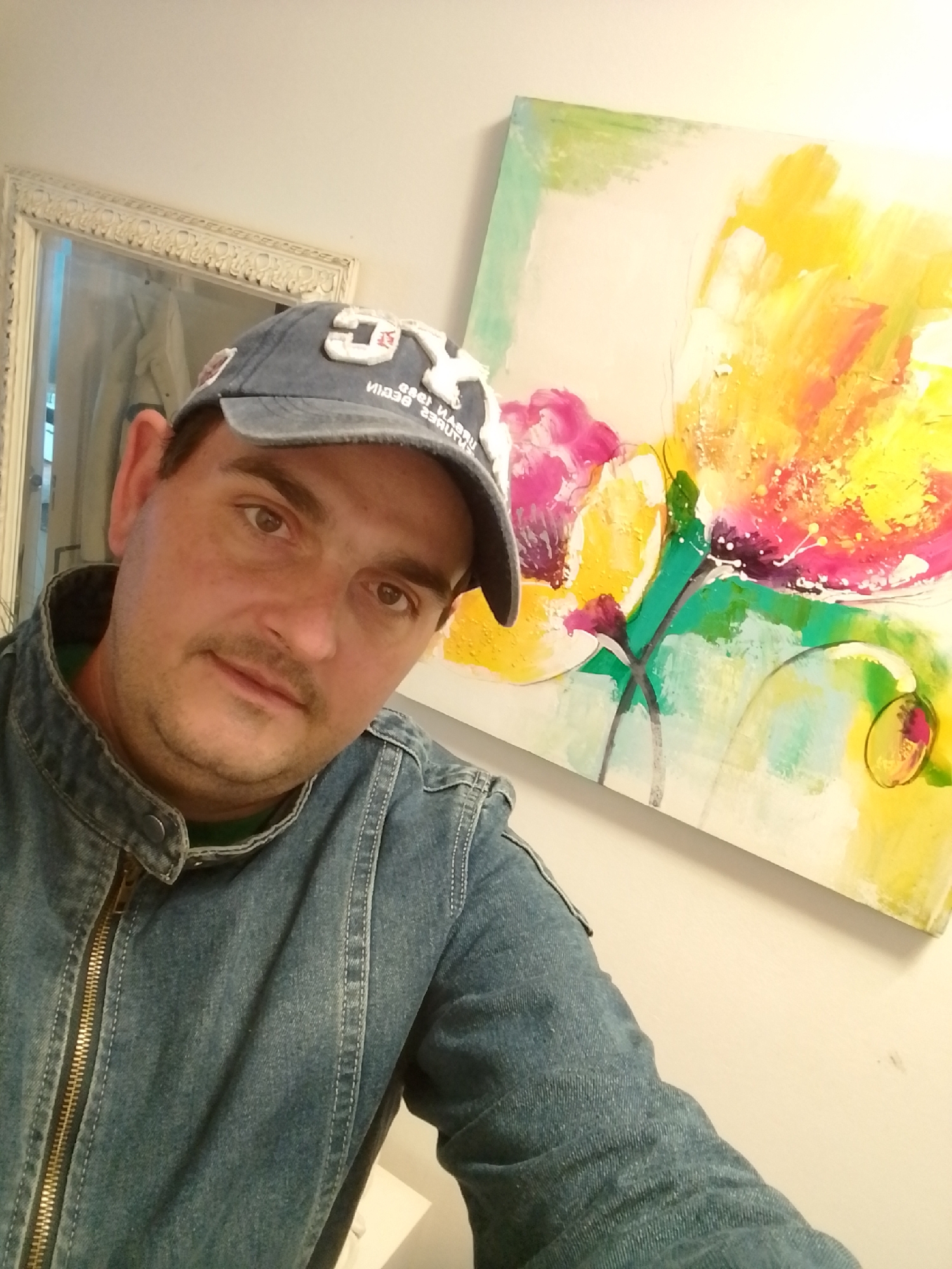 A medium shot captures a man wearing a denim jacket and a blue baseball cap. He is looking directly at the camera with a neutral expression. The cap has white lettering. In the background, a large, abstract painting with vibrant colors, primarily yellow, pink, and green, hangs on a light-colored wall. To the left, a portion of an ornate, white-framed mirror is visible, reflecting a dimly lit interior. The lighting appears to be artificial, casting a slight shadow on the man's face. The setting is indoors, likely a residential or gallery space. The exact location in the city of Wedel, Germany, is not discernible from the image.