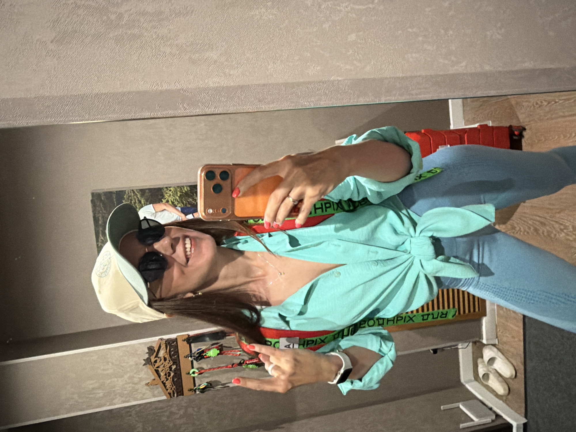 A woman takes a mirror selfie indoors. She is wearing a light blue button-down shirt tied at the waist, light blue leggings, a light-colored baseball cap, and sunglasses. She is also wearing a red backpack with green straps. The woman is smiling and giving a rock-and-roll hand gesture with her left hand. She is holding an orange smartphone in her right hand. In the background, there is a mirror, a wall with textured wallpaper, a key holder with several keys, a red suitcase, and a pair of white slippers. There is also a framed picture on the wall. The overall environment suggests a domestic or indoor space. The location in Birobidzhan, Russia, is not discernible from the visual elements.