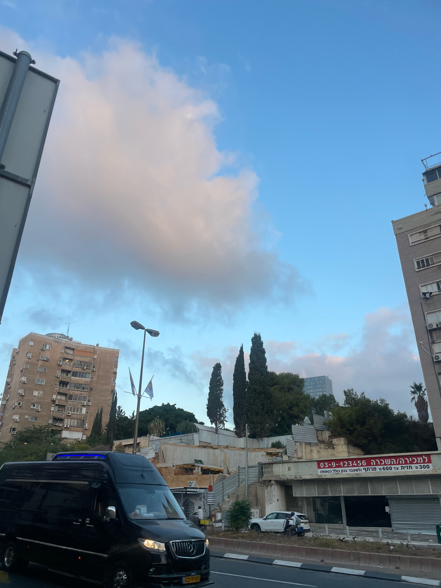 In Ramat Gan, Israel, a dark, four-door van with blue emergency lights on its roof is parked on the right side of a multi-lane road. The van has tinted windows and a visible license plate. A white SUV is parked behind the van, with two individuals standing next to it. In the background, a multi-story building with many windows and air conditioning units is visible, along with several tall, dark green trees. A construction site with scaffolding and debris is also present. Overhead, a blue sky is dotted with fluffy white clouds. A street lamp with two lights stands near the building and trees. A white sign is visible on the right side of the road with Hebrew writing and a phone number.