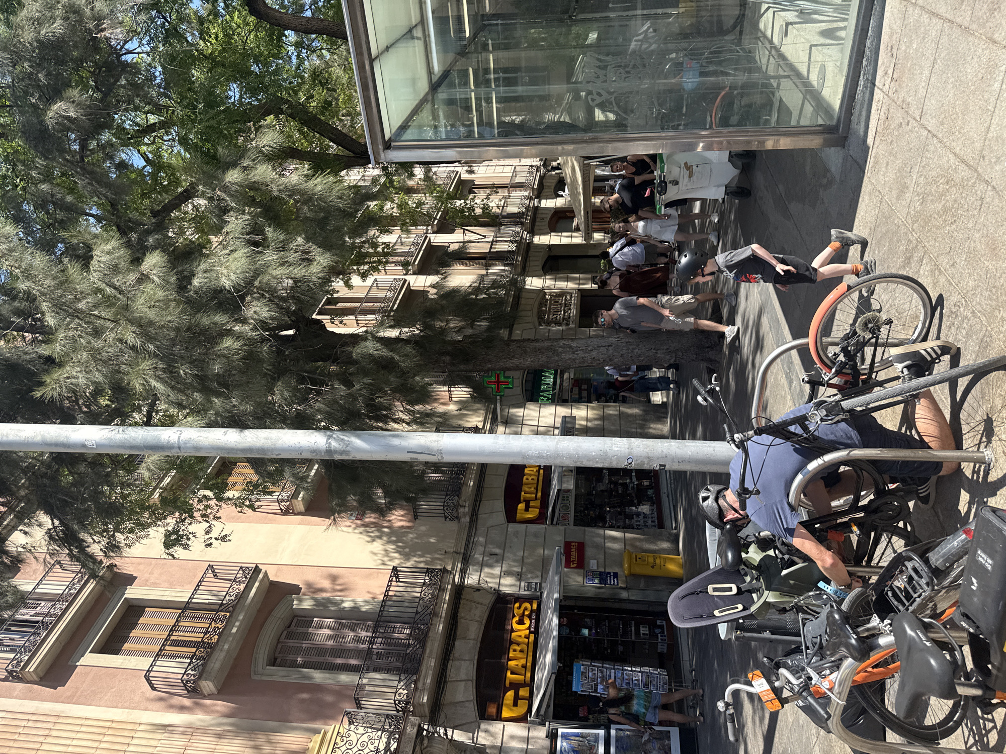 The image depicts a sunny street scene in Barcelona, Spain, likely in the Gothic Quarter or Eixample district, given the architectural style. In the foreground, a man wearing a blue t-shirt and black shorts, with a helmet on his head, is kneeling and appears to be locking a bicycle. Several other bicycles are parked nearby, including an orange one in the lower left. To the right of the man, a young boy in a grey t-shirt and black shorts walks past a modern glass structure, which might be an entrance to an underground facility or a modern bus stop.
In the background, people are walking on the sidewalk in front of buildings with a distinctive architectural style featuring stone facades, balconies with ornate railings, and shuttered windows. Several storefronts are visible, including "TABACS" (tobacco shops) and a "FARMACIA" with a green cross symbol. A large tree with dense foliage provides shade over part of the street. The overall impression is of a bustling urban environment on a bright day.