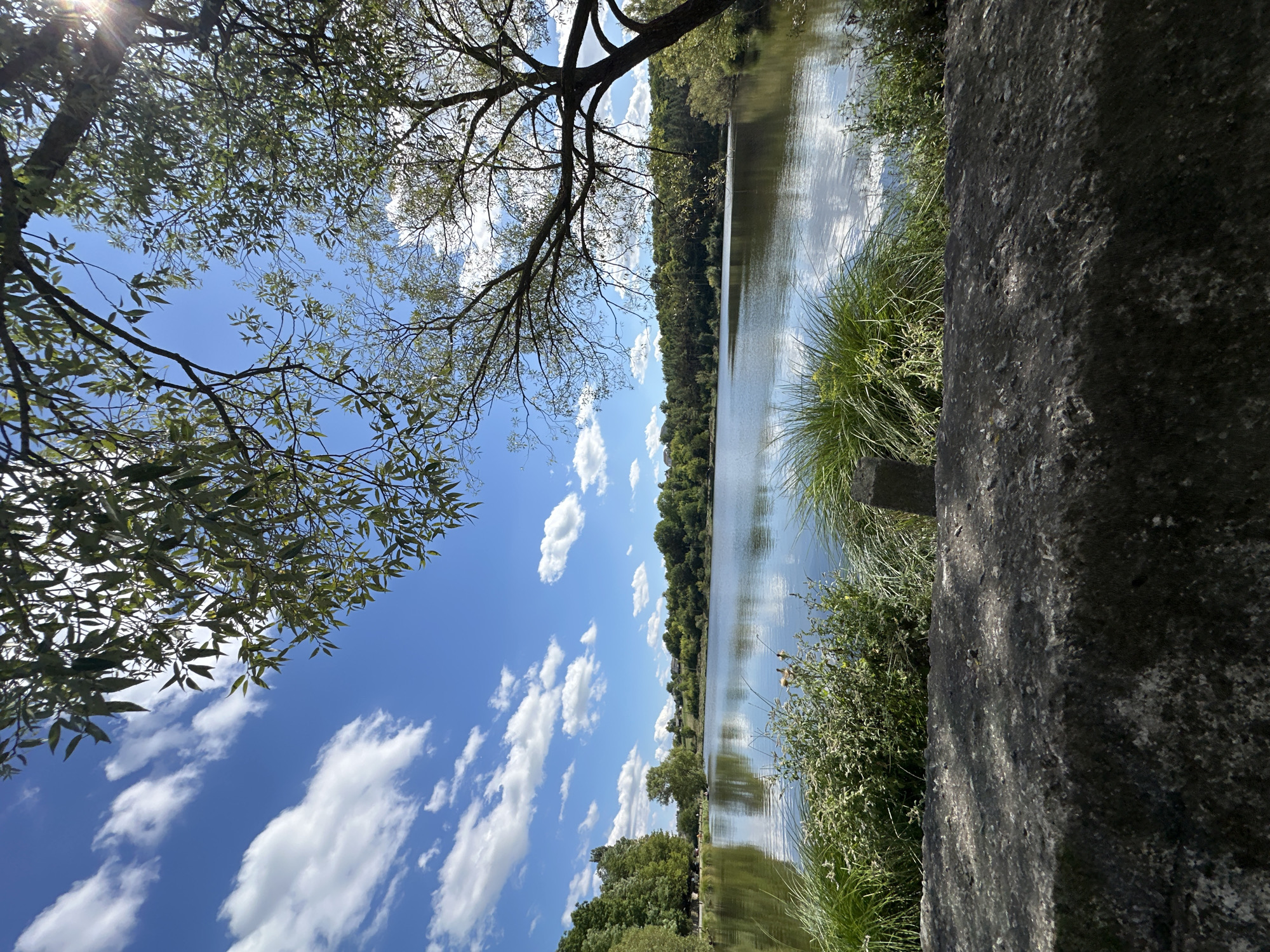 The image depicts a serene lake scene, likely situated within the city of Makiv, Ukraine. The foreground is dominated by a rough, textured stone wall, appearing weathered and partially shaded. Beyond the wall, lush green grasses and vegetation grow along the water's edge. The lake itself is calm, reflecting the bright blue sky dotted with fluffy white clouds. A line of trees forms a dense backdrop along the far shore, interspersed with some buildings visible in the distance. The sun's rays are breaking through the branches of a large tree on the right side of the frame, casting dappled light and a lens flare effect. No people or animals are clearly visible in the frame. The overall impression is one of natural tranquility.