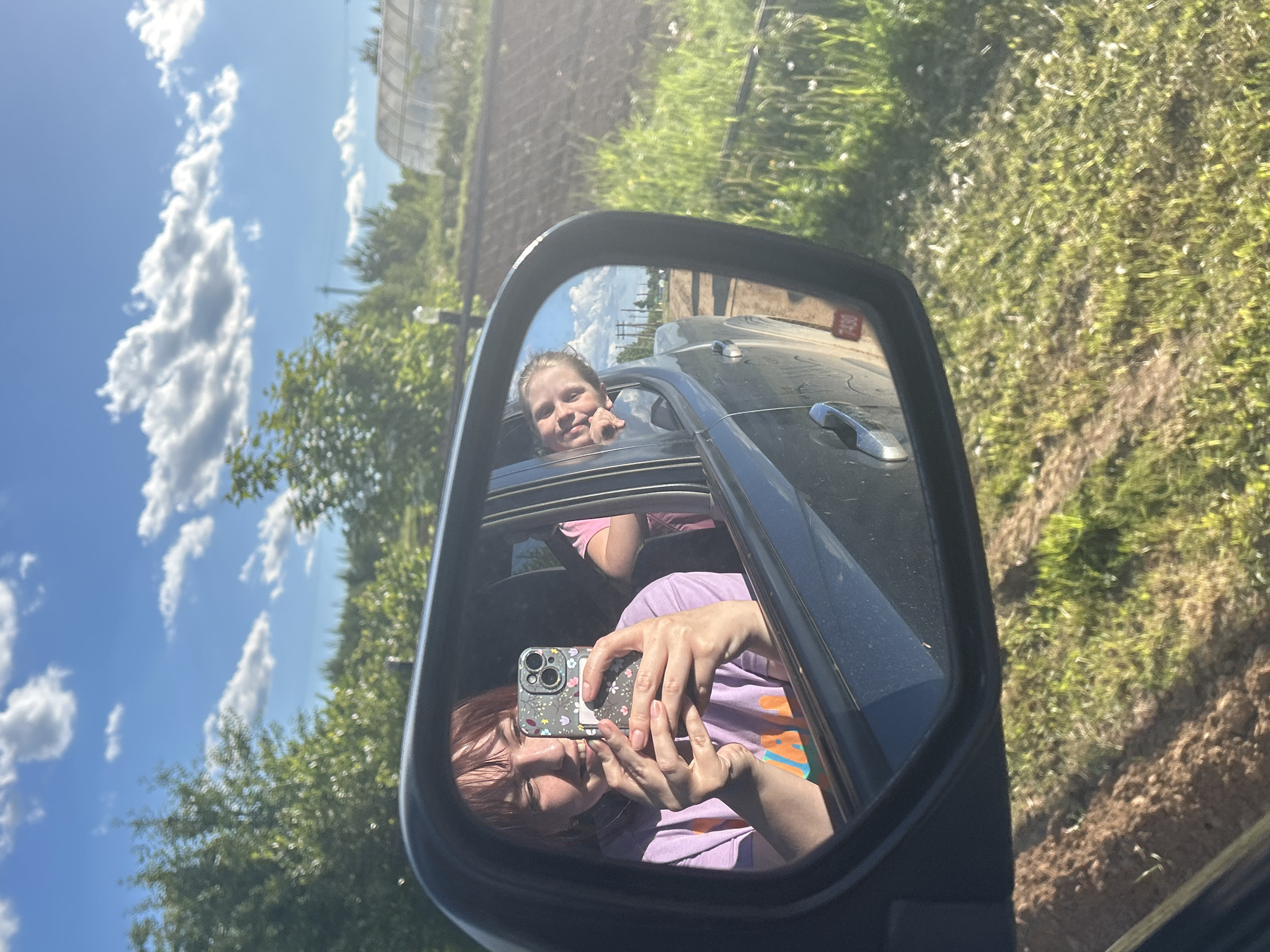 The image shows a view from the side mirror of a car. In the mirror's reflection, there are two individuals: an adult with reddish hair wearing a purple shirt and holding a smartphone with a flowered case, apparently taking a photo, and a young girl in a pink shirt looking out the car window. The car is black. The background outside the car shows a blue sky with white clouds, trees, grass, and what appears to be a greenhouse. The location is Фоки, Russia.